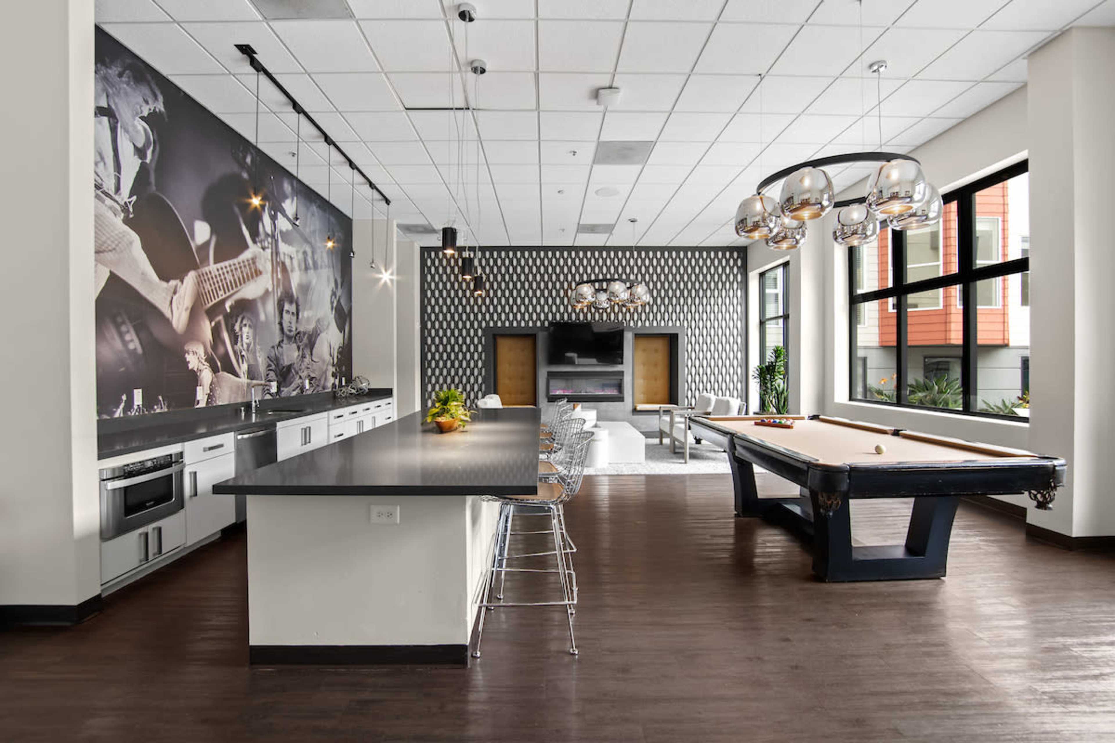 The image shows a modern common area featuring a kitchen bar with stools, a pool table, and large windows with a striking black-and-white mural on one wall.