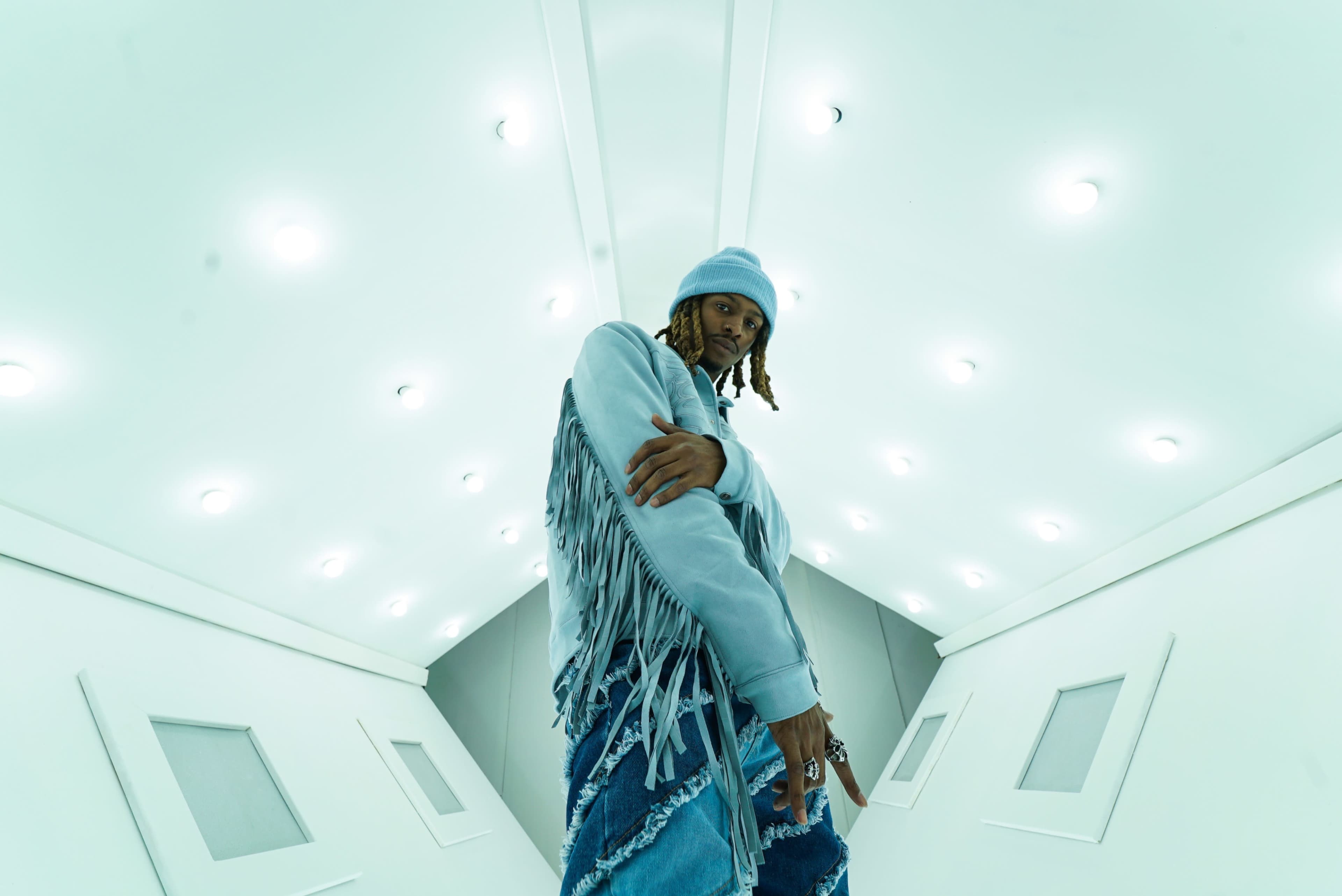 A person with braided hair wearing a fringed jacket and denim jeans poses in a well-lit room with white walls and a geometric design.