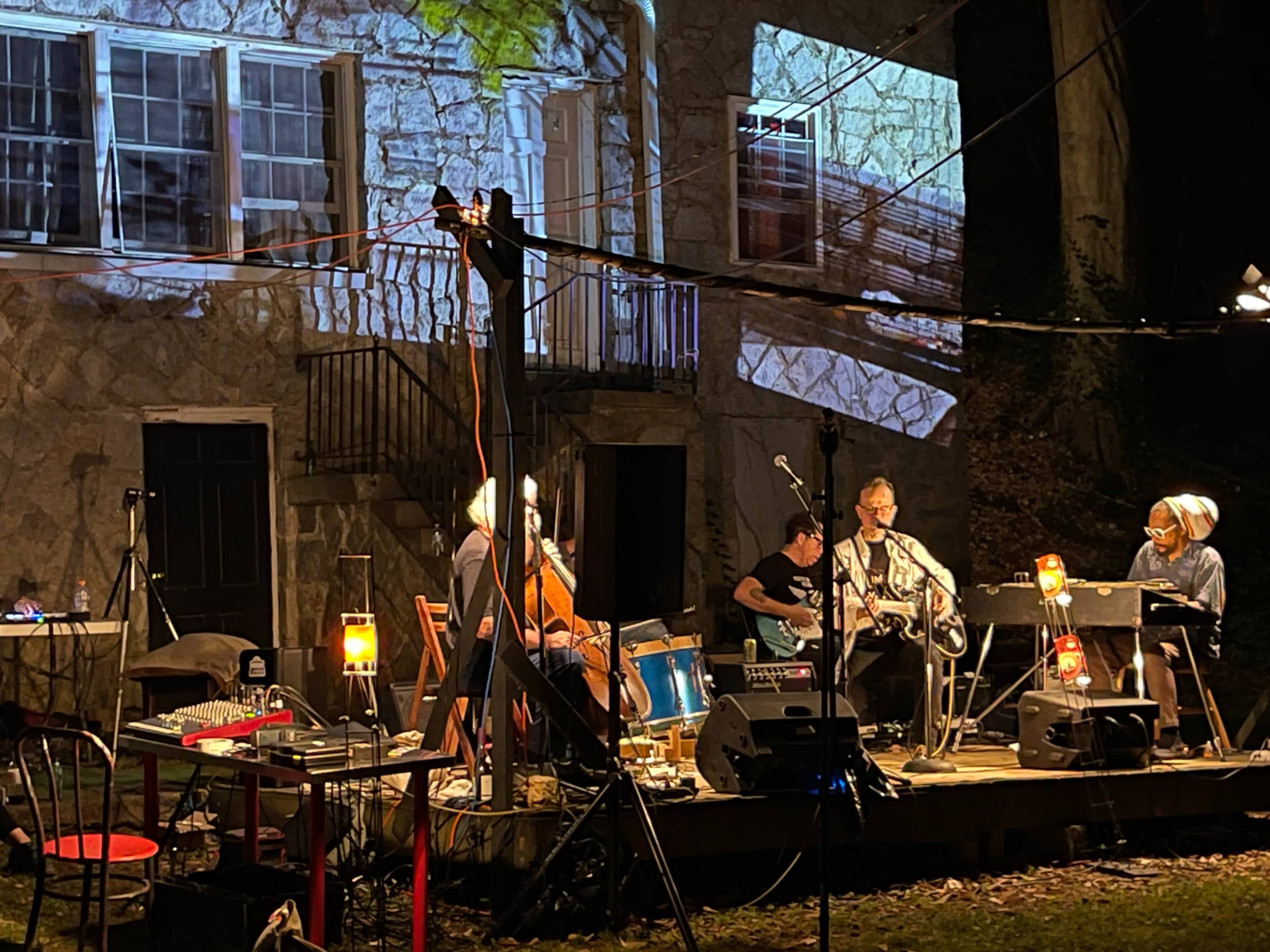 A small outdoor music performance takes place in front of a stone house, with musicians playing instruments and visuals projected onto the house's exterior.
