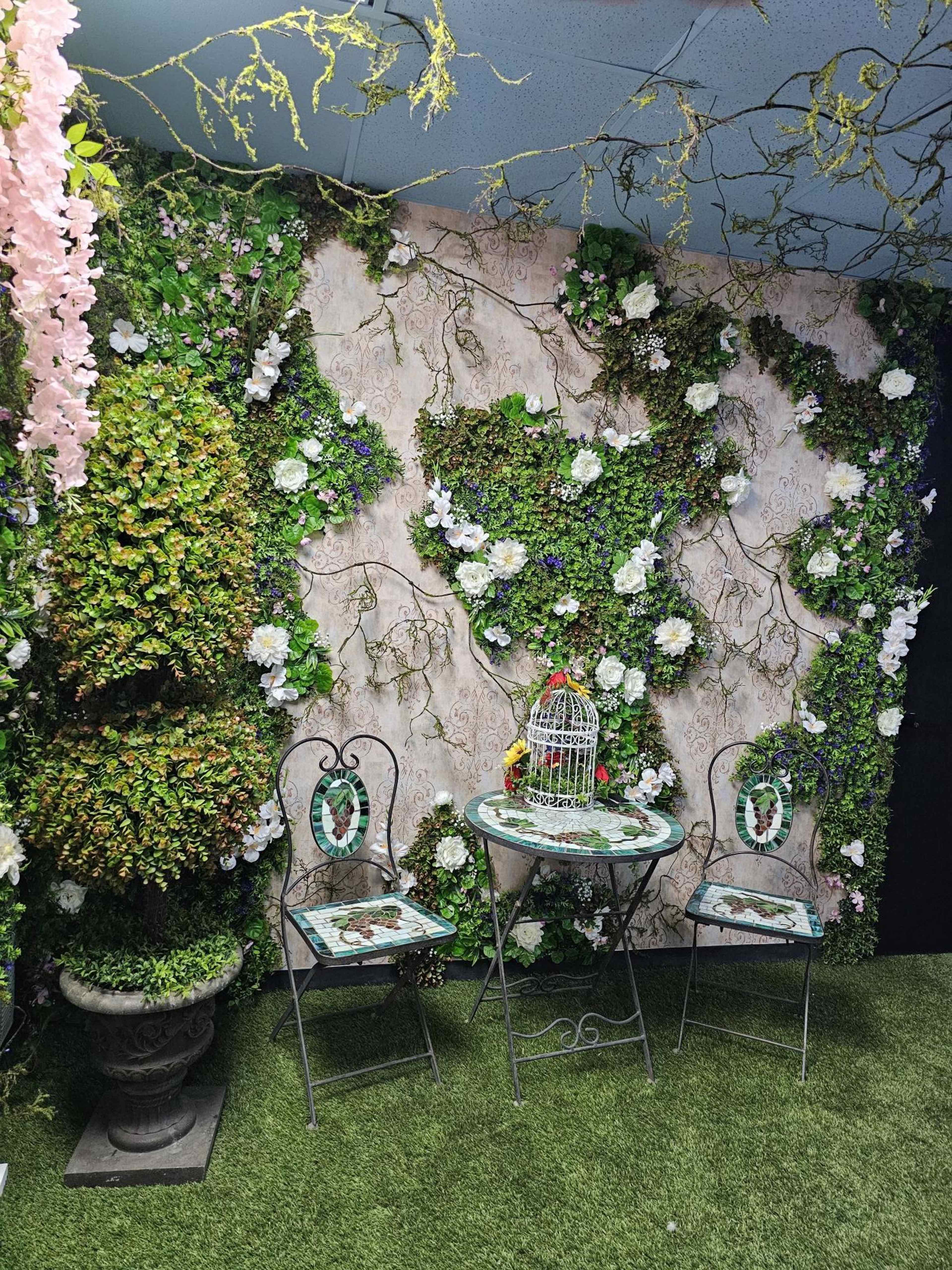 A decorative indoor space featuring a wall covered in greenery and flowers, with a small round table and two chairs set in front of it, alongside a birdcage.