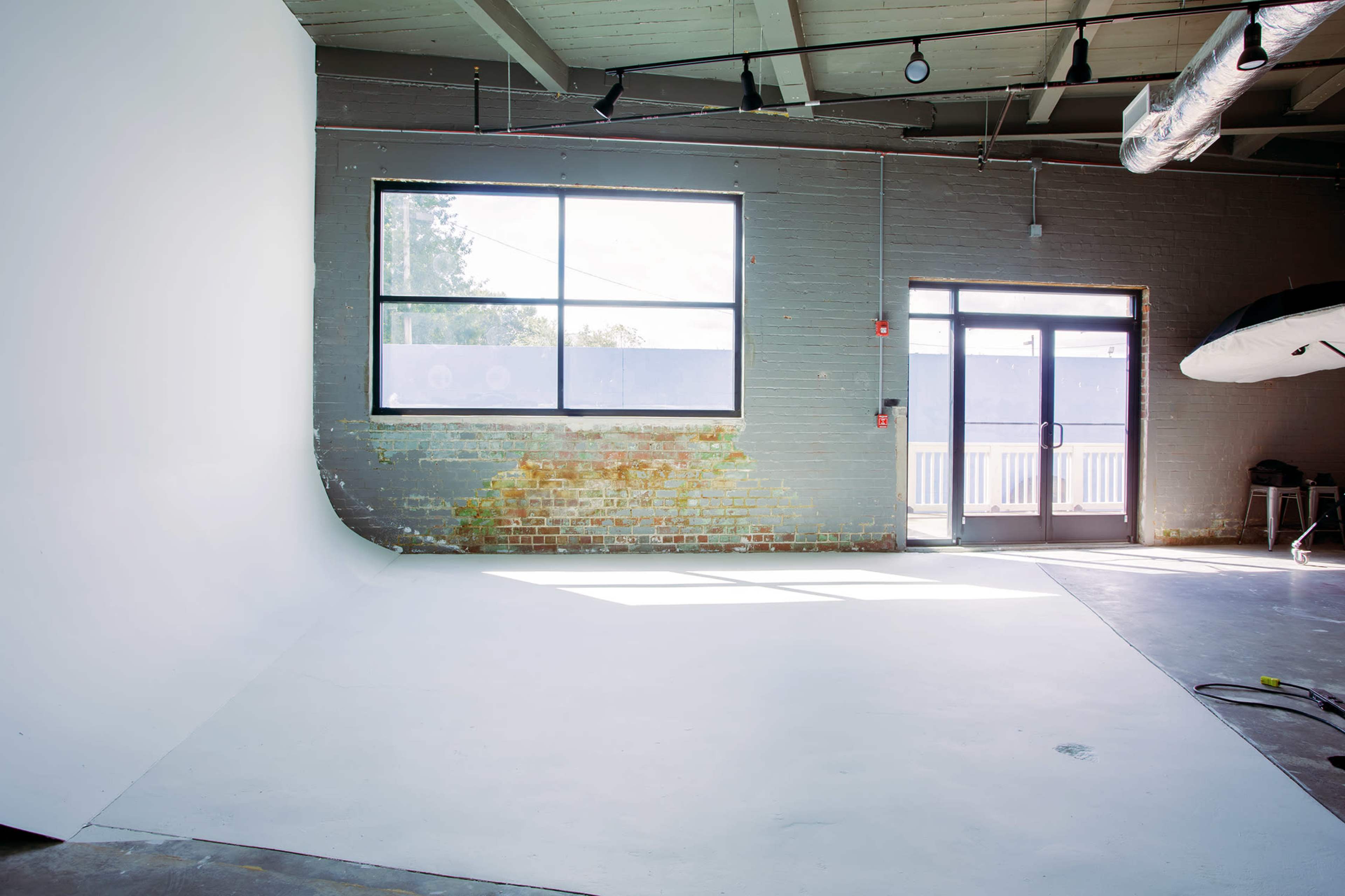 The image shows a spacious interior with a white curved wall, a large window, and a glass door, set against a backdrop of exposed brick.