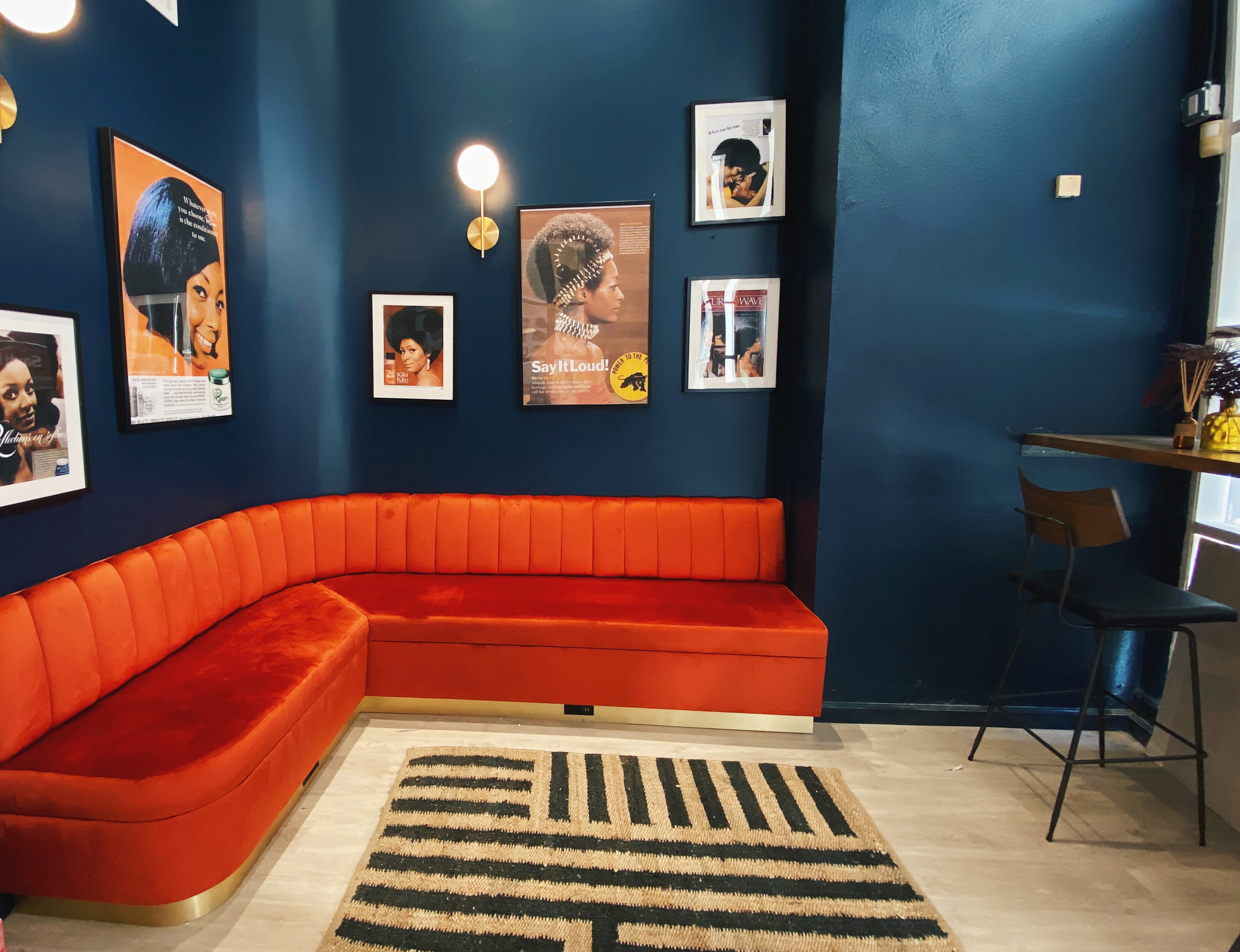 A cozy interior of a lounge featuring an orange velvet curved sofa, framed posters on the dark blue walls, and a small wooden bar table with chairs.