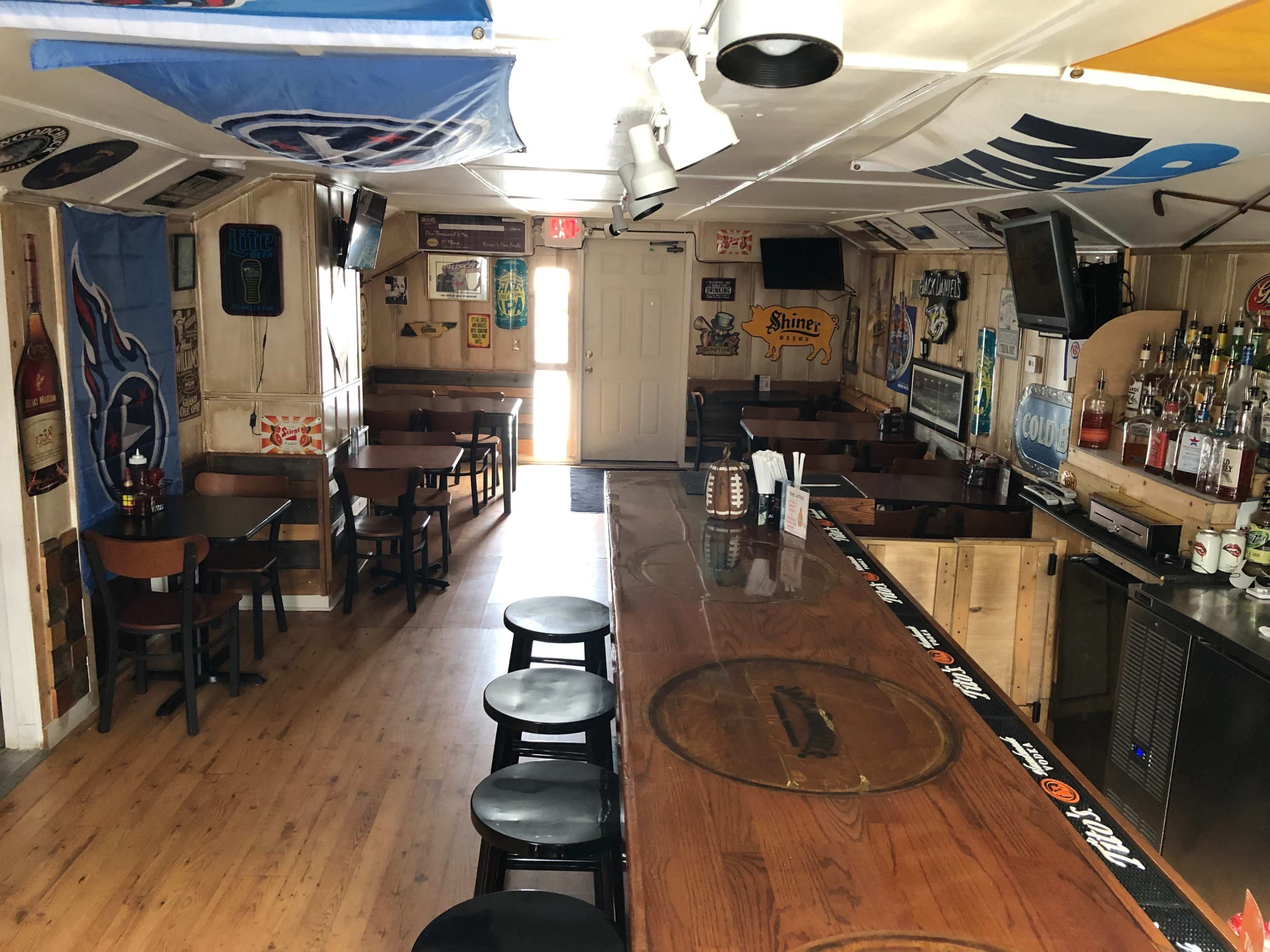 The interior of a bar with a long wooden counter and several empty tables and chairs, adorned with various flags and decorations on the walls.