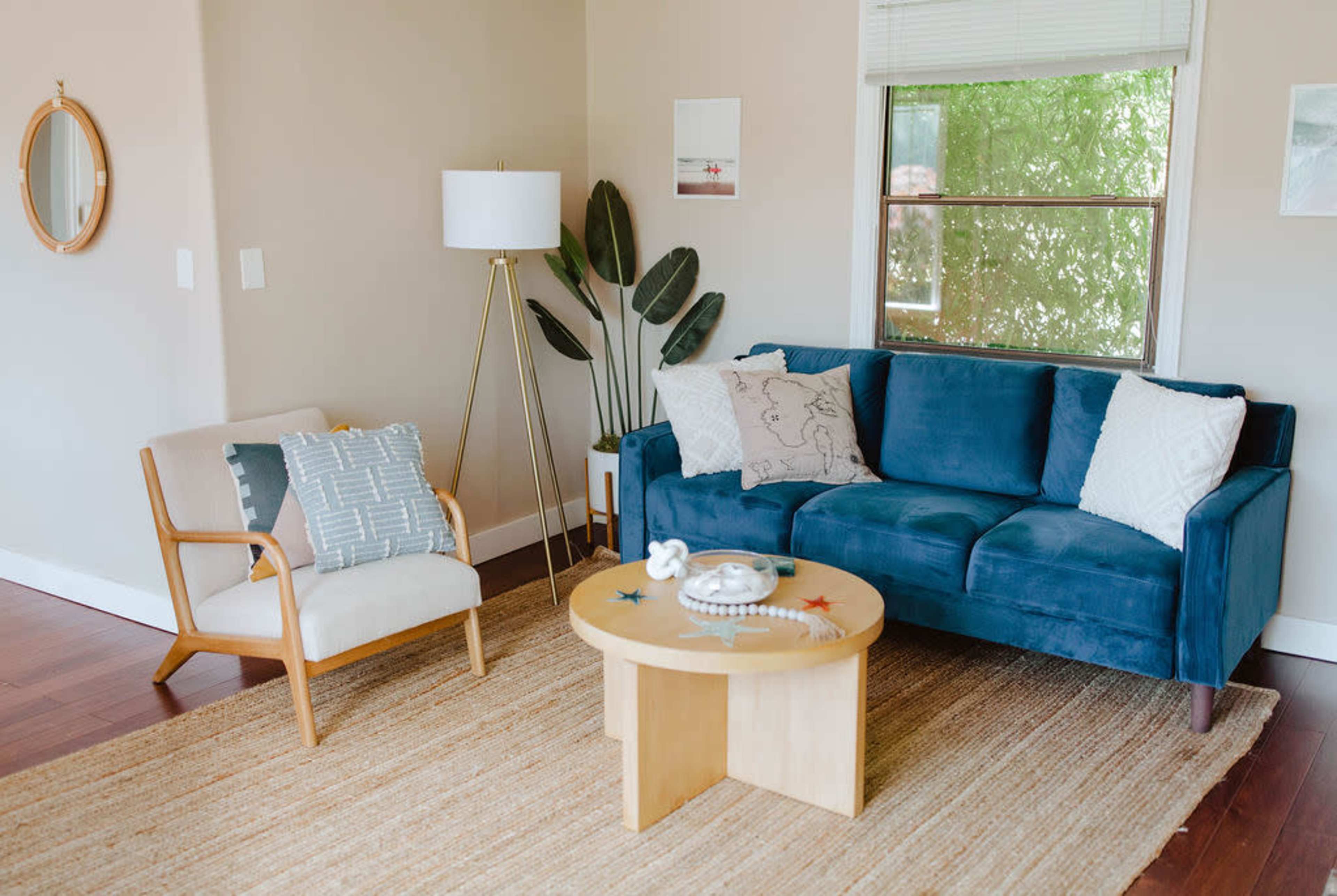 A living room featuring a blue sofa, a light-colored armchair, a round coffee table, and a floor lamp, with plants and framed pictures on the walls.