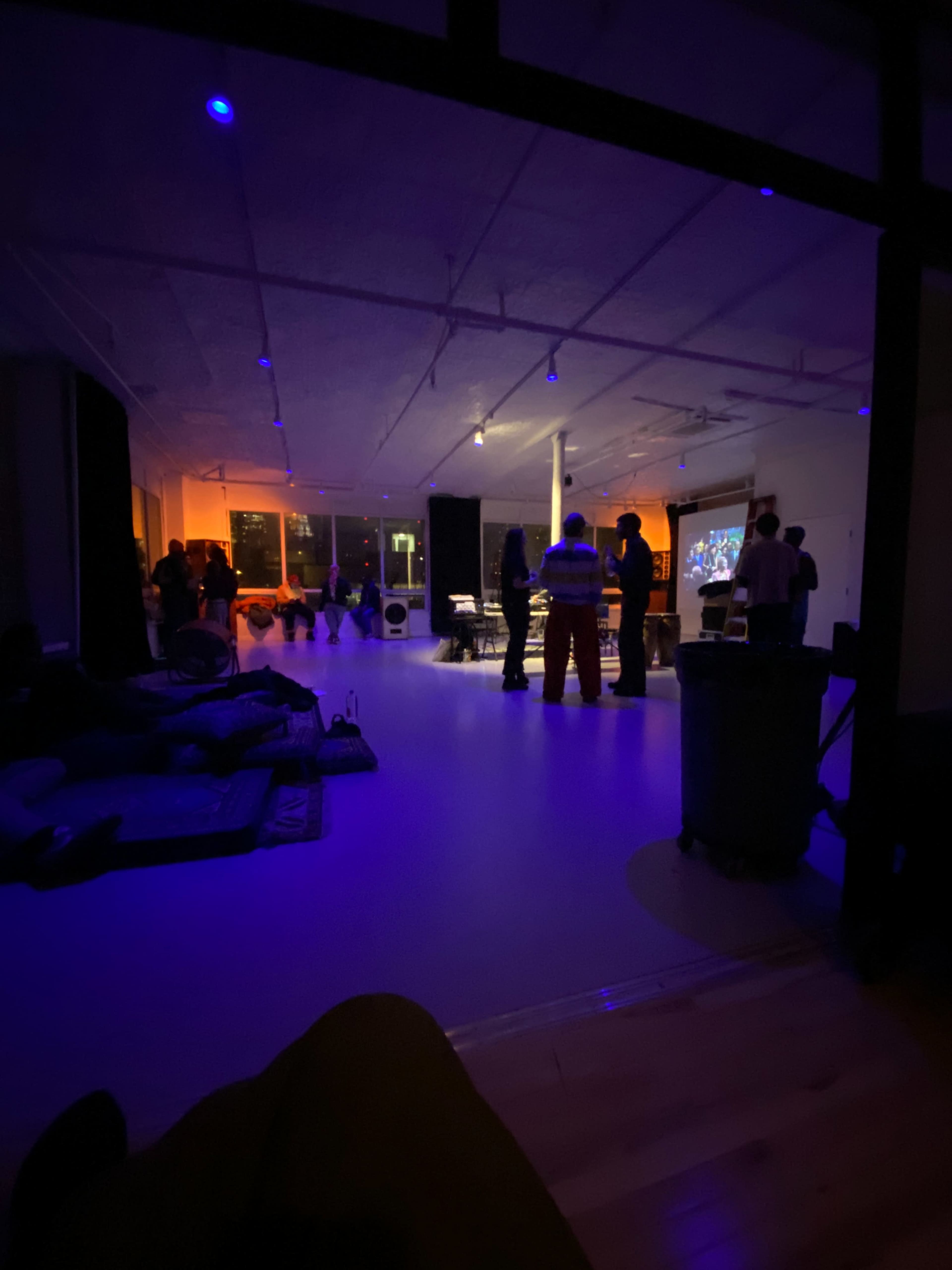 A group of people gathers in a dimly lit room with colorful lights and a large screen displaying images.