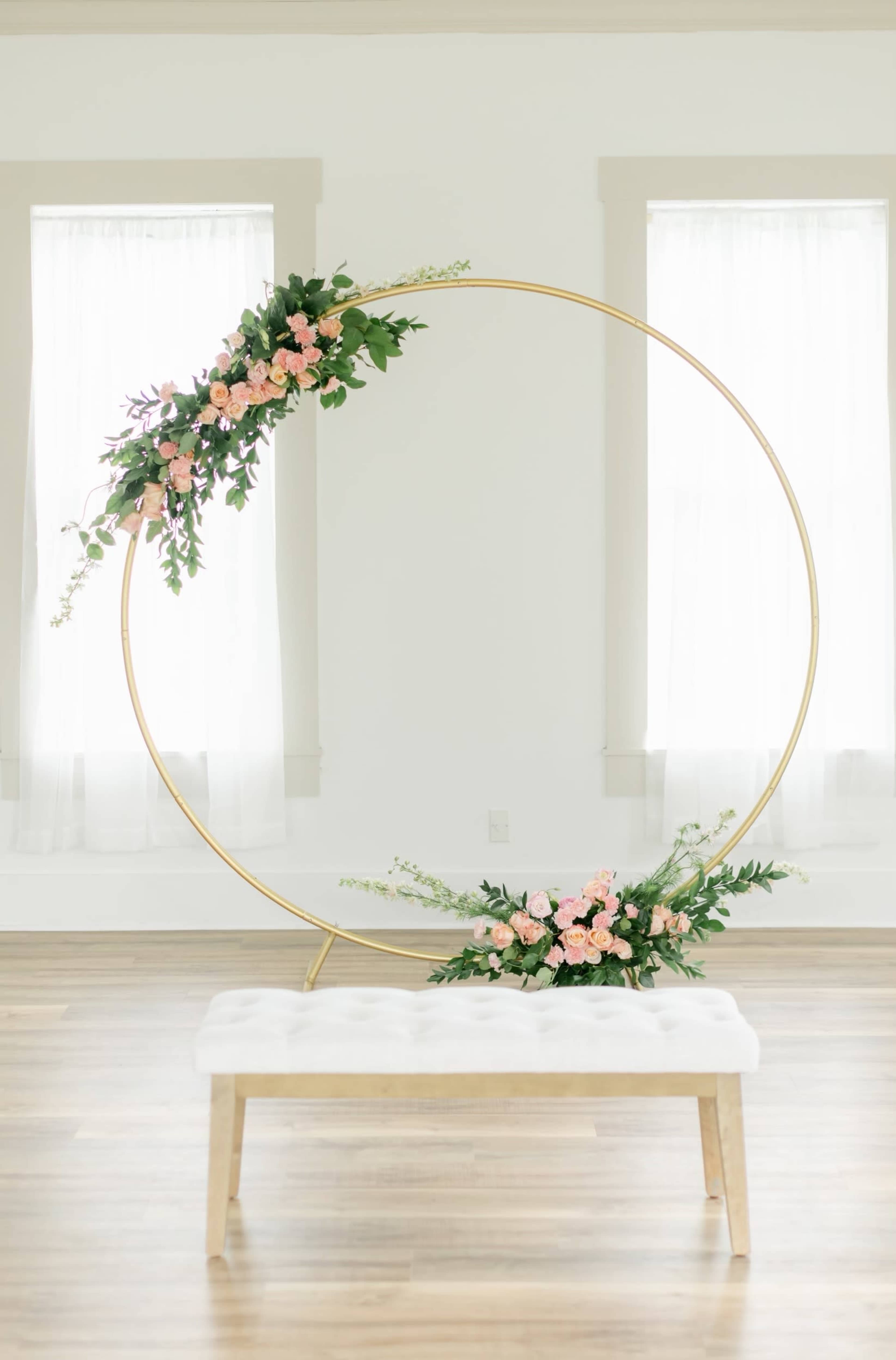 A circular gold frame adorned with pink flowers is positioned beside a white bench in a room with large windows.