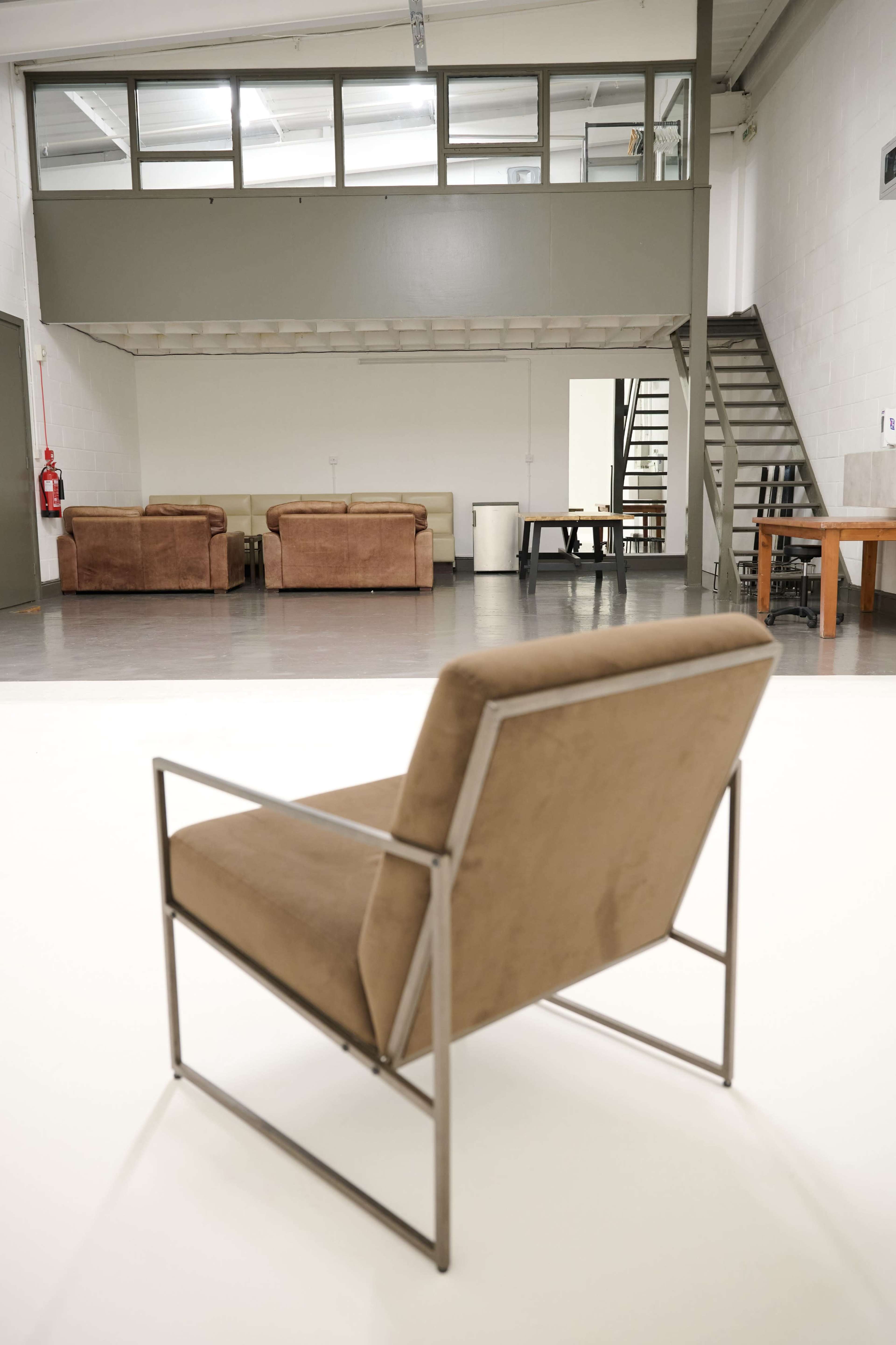 A modern space features a brown upholstered chair in the foreground, with two sofas, a table, and a staircase visible in the background.