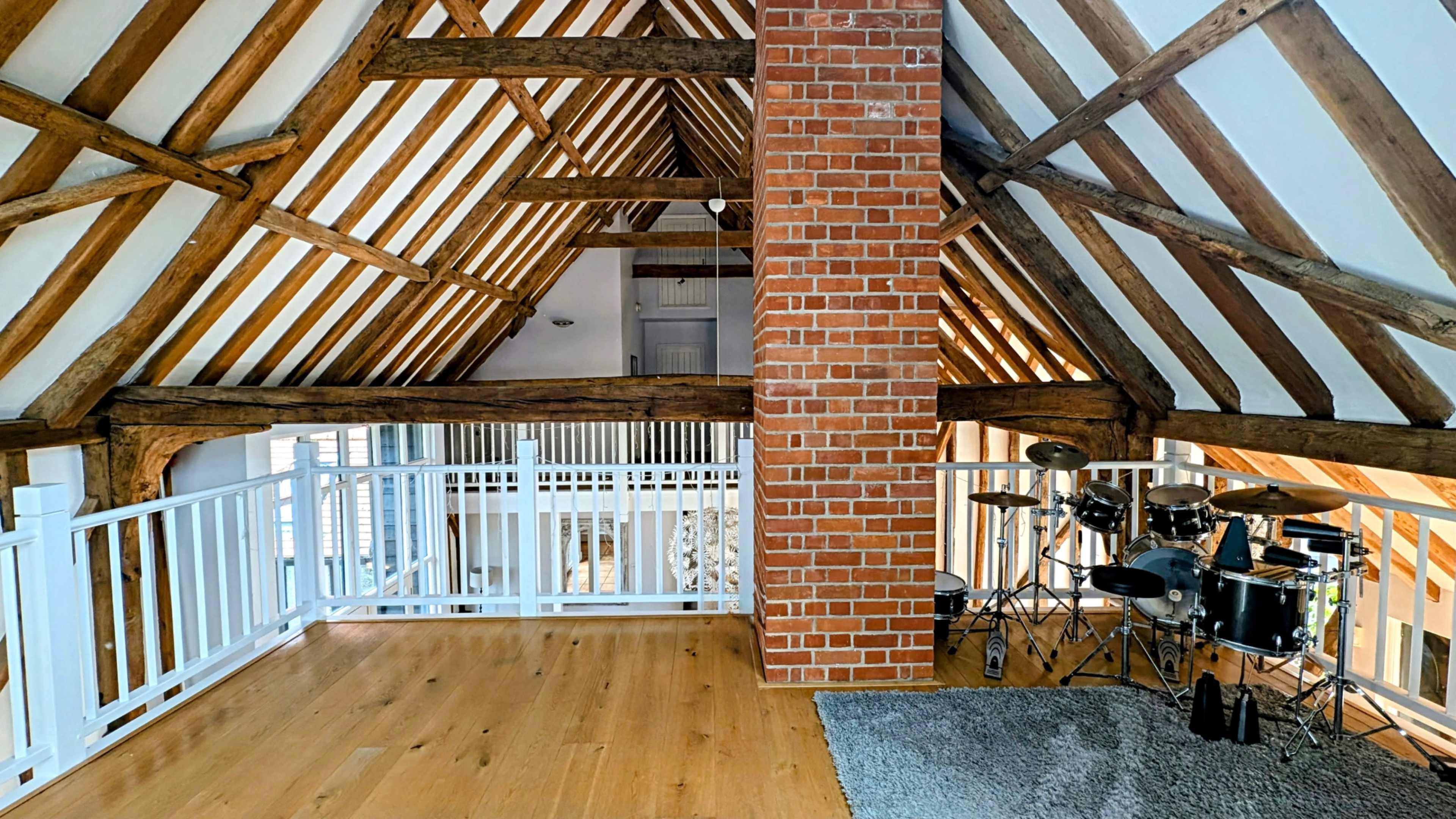 The image shows an interior space with wooden beams, a brick column, and a small drum set positioned beside a white railing.