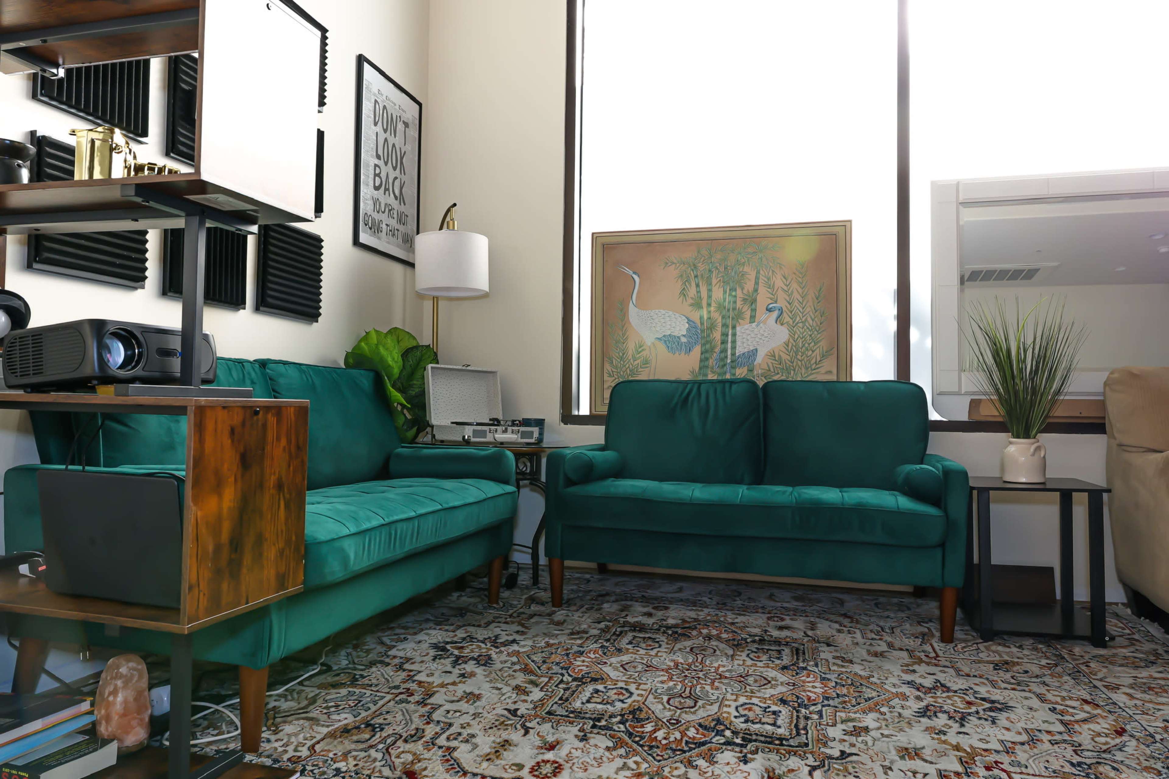The image shows a living room corner with two teal sofas, a wooden side table, a projector, wall art, and a large window.