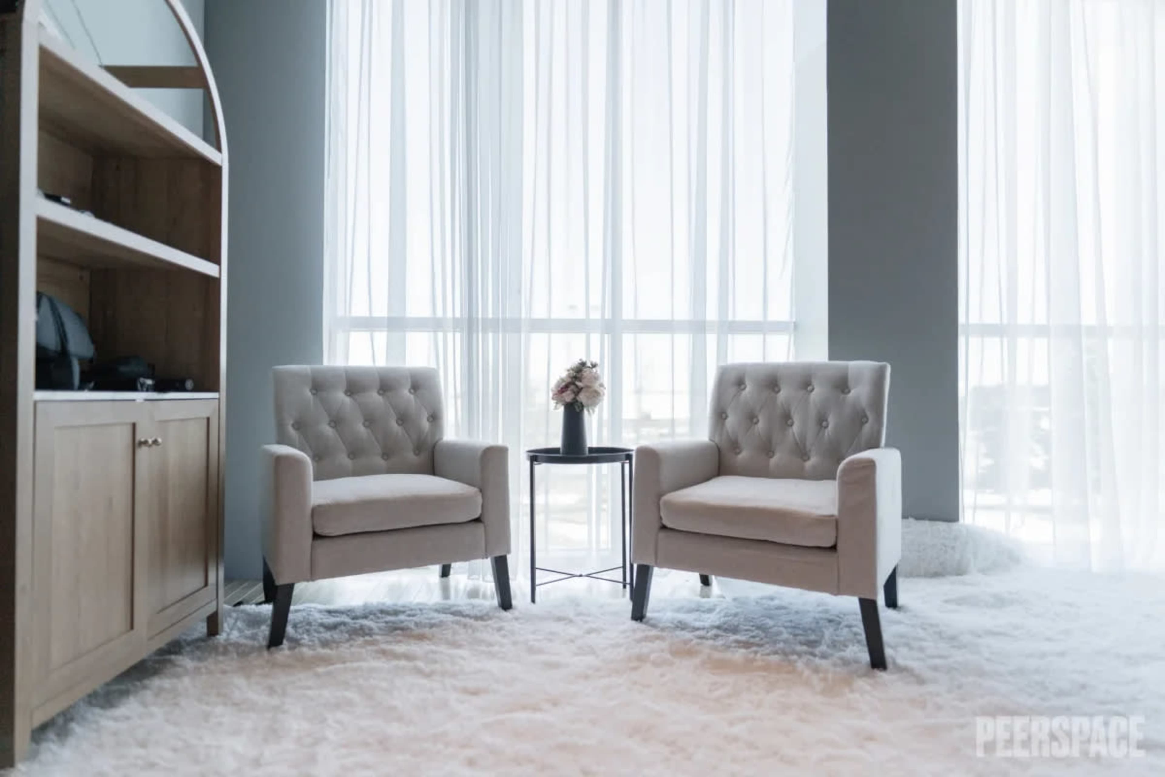 Two tufted armchairs are positioned beside a small round table in a bright room with large windows draped in sheer curtains.