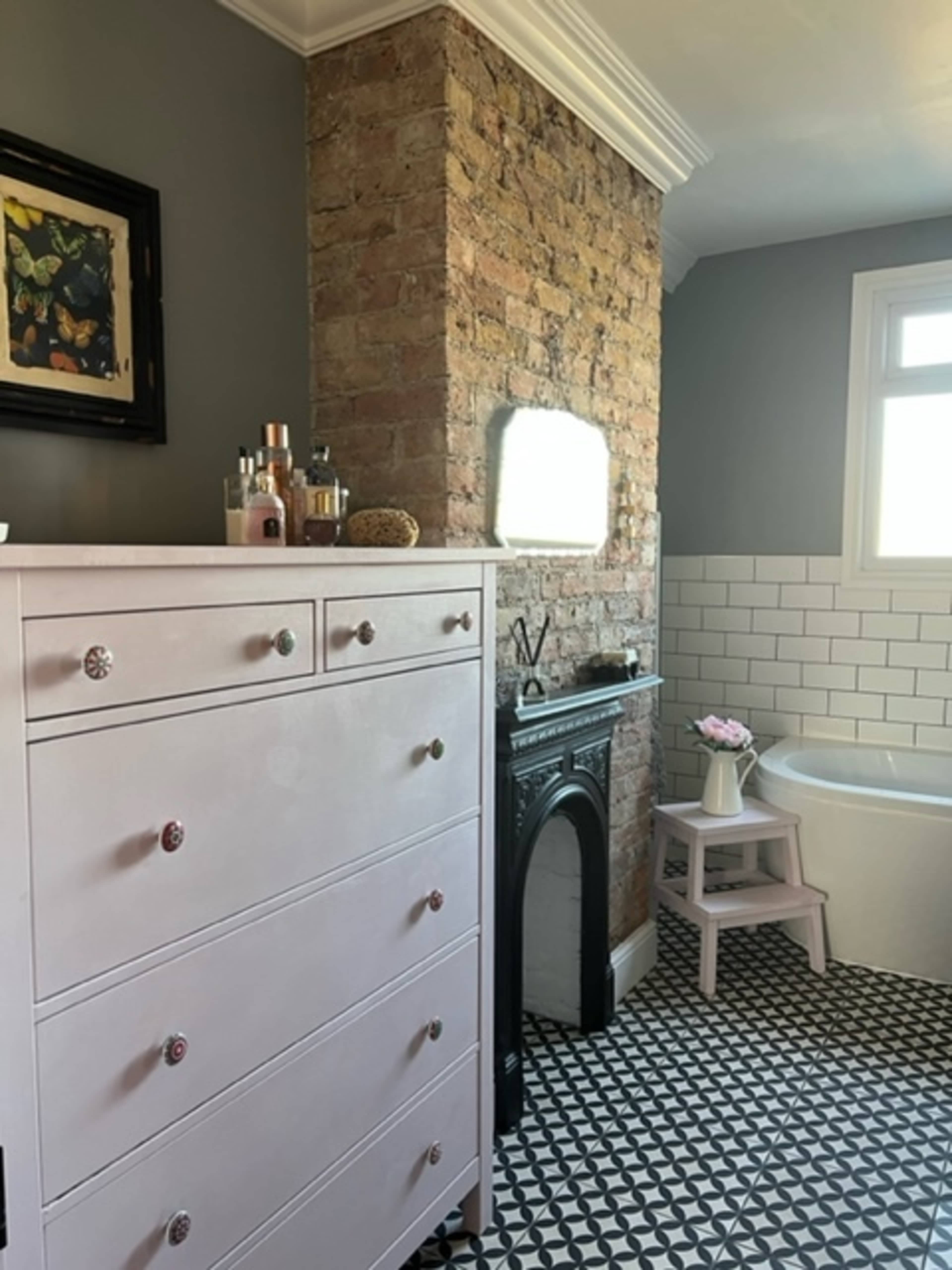 A modern bathroom features a pink dresser, exposed brick wall, vintage fireplace, and a freestanding bathtub, with a patterned tiled floor and a window allowing natural light.