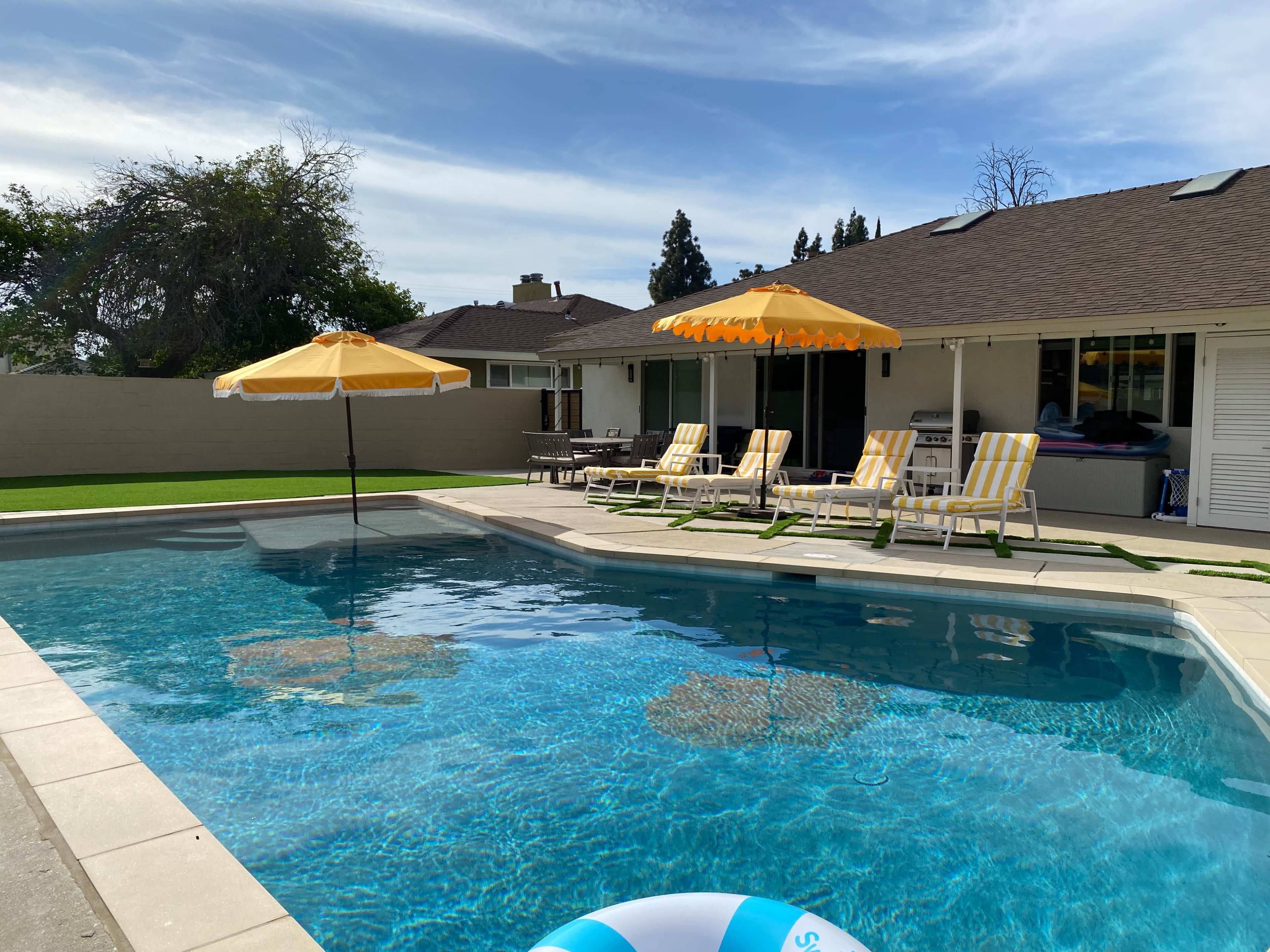 A residential backyard with a swimming pool, two yellow umbrellas over lounge chairs, and a house in the background.