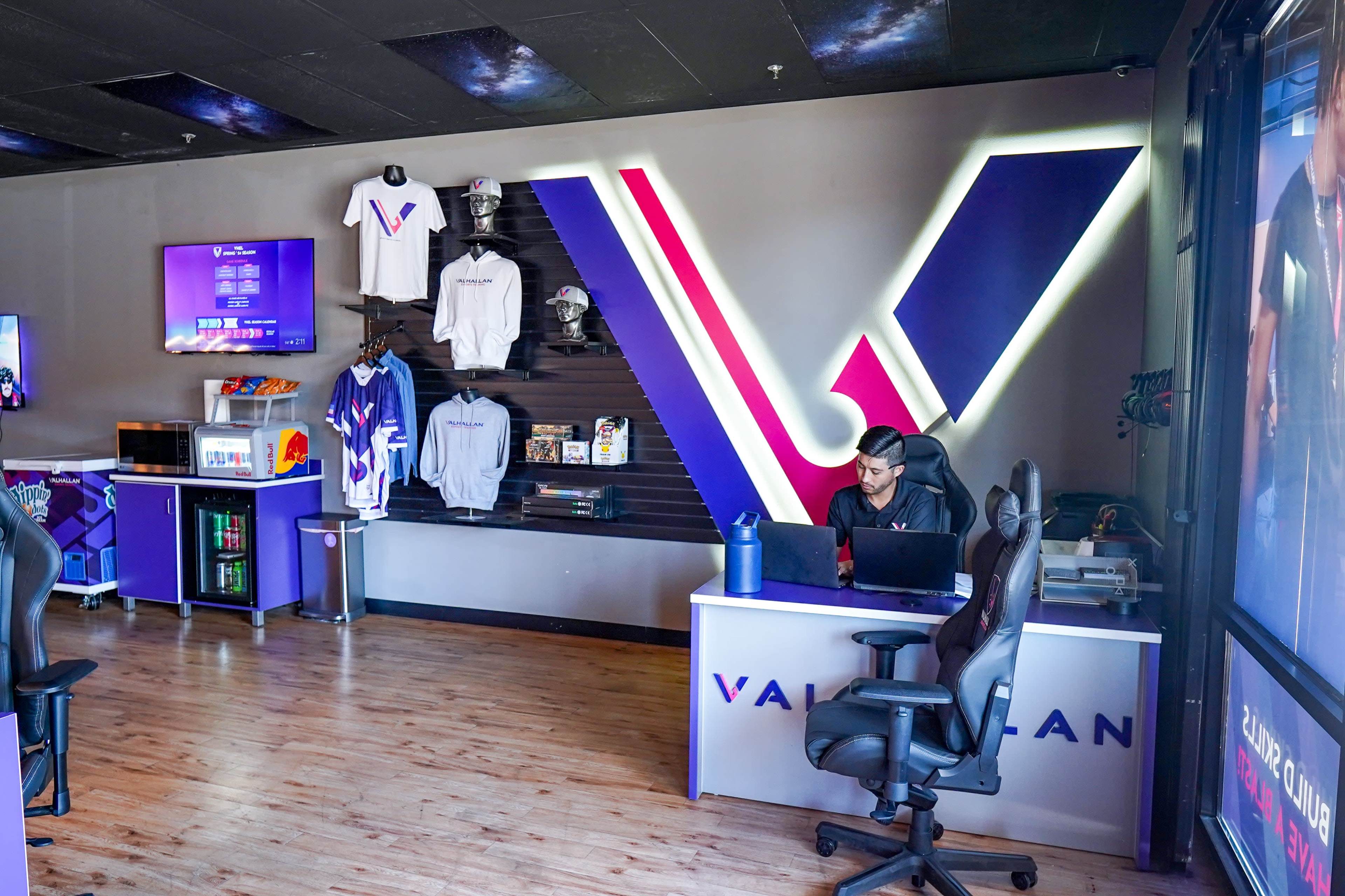 The image shows a modern gaming lounge with promotional merchandise displayed on the wall, and a person seated at a desk working on a laptop.
