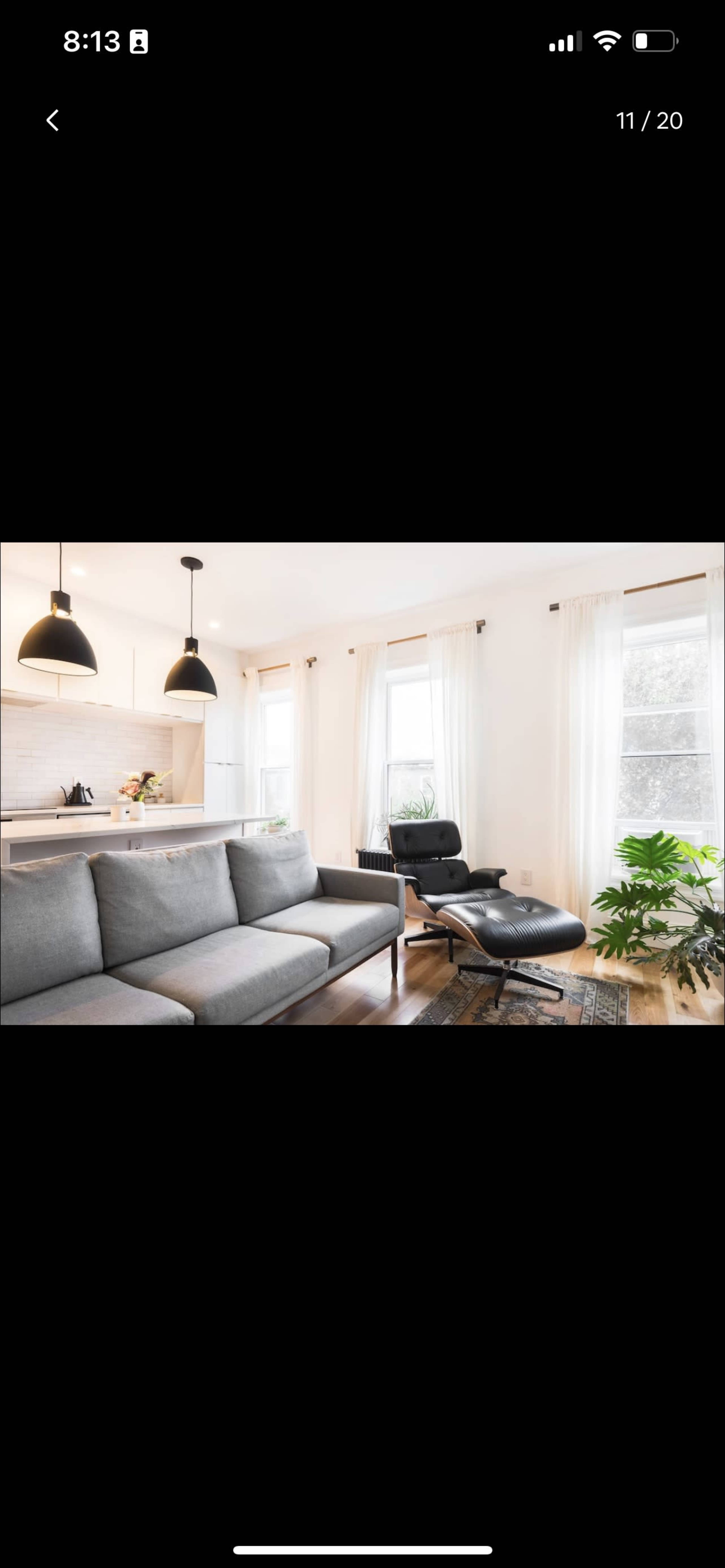 The image shows a modern living room with a gray couch, an ergonomic chair, large windows with sheer curtains, and a potted plant.