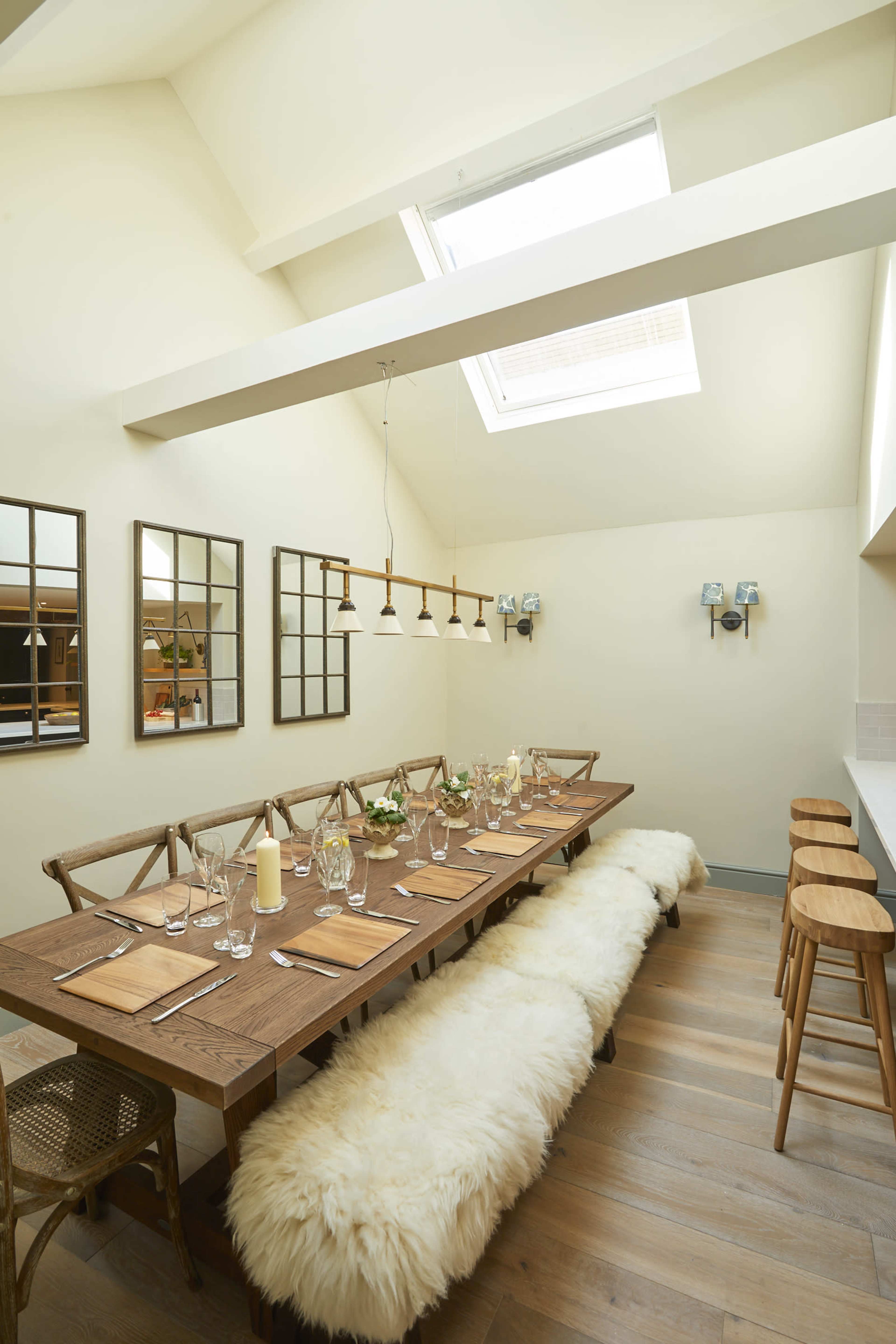 A well-lit dining room featuring a long wooden table with set place settings, surrounded by wooden chairs and a soft bench, under a skylight and framed mirrors on the walls.