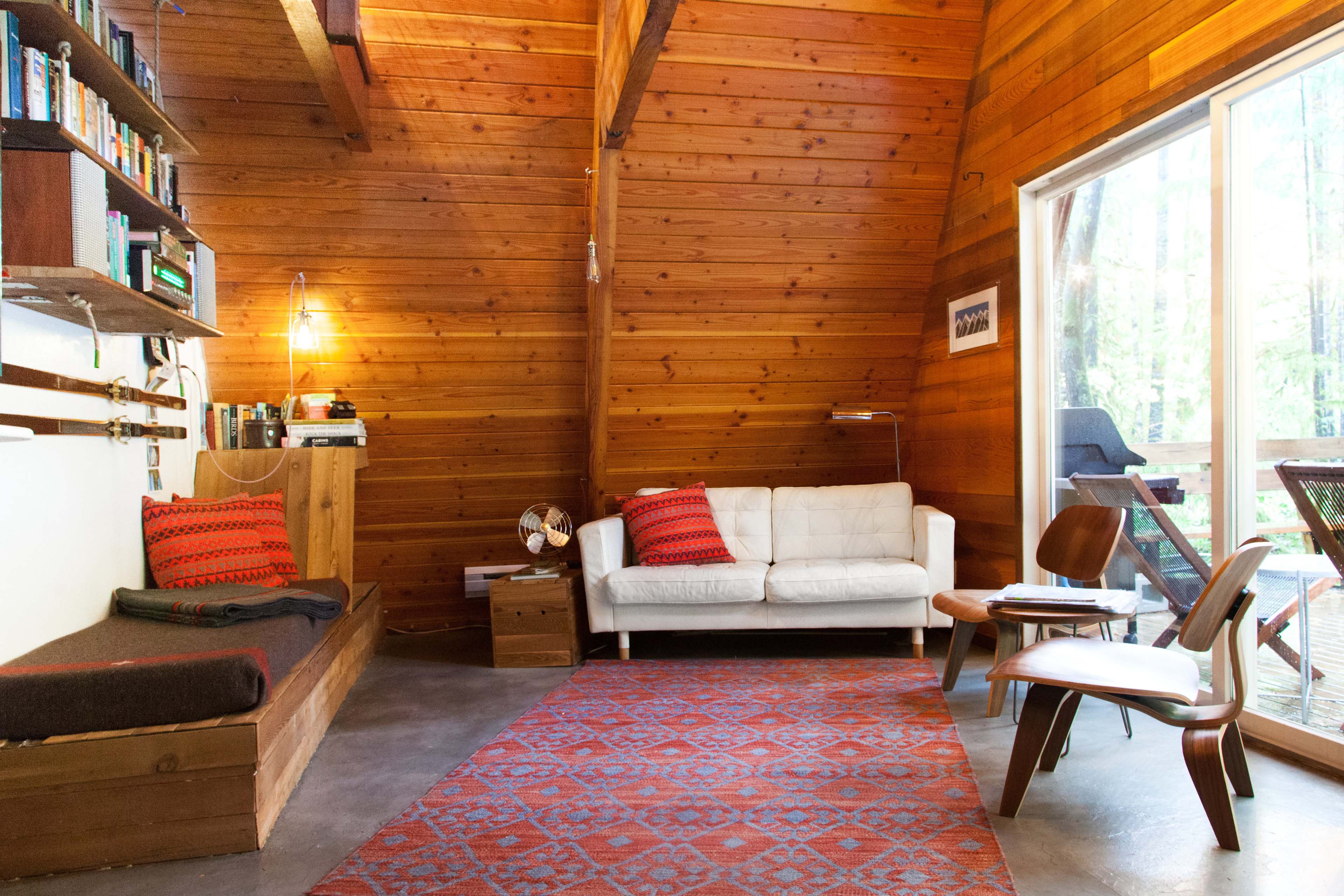 A cozy living room with wooden walls, a white couch, a red-patterned rug, and large windows offering a view of the outdoors.