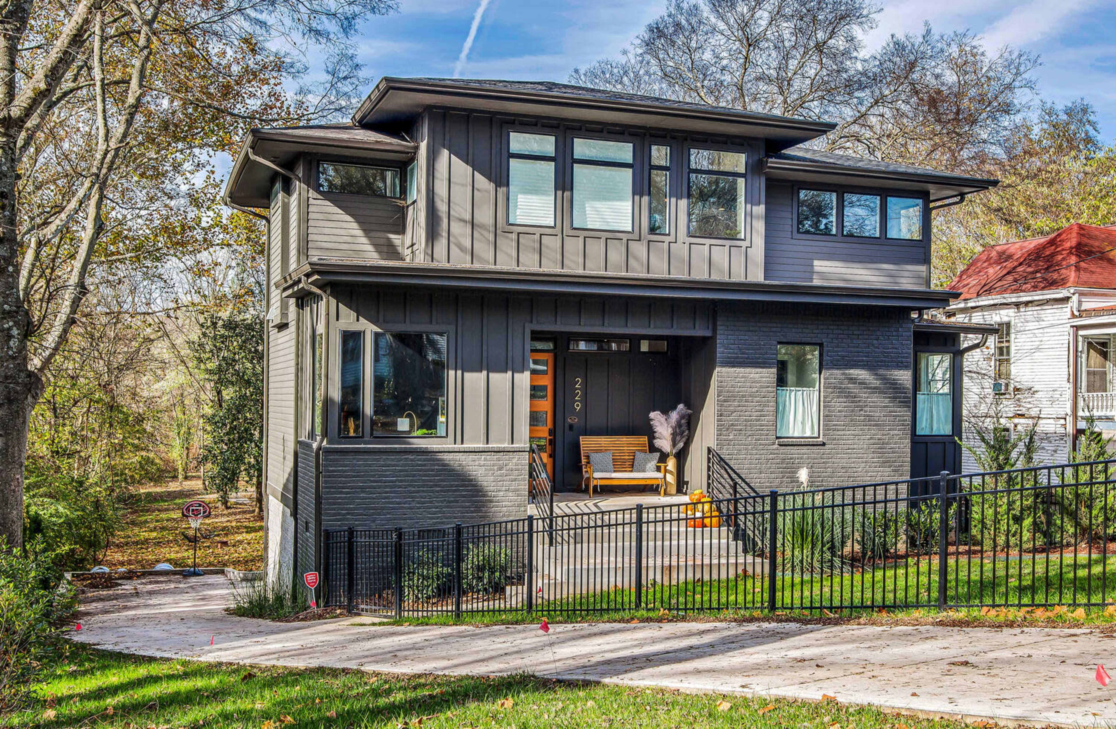 The image shows a modern two-story gray house with large windows, a porch, and a black iron fence in a landscaped yard.