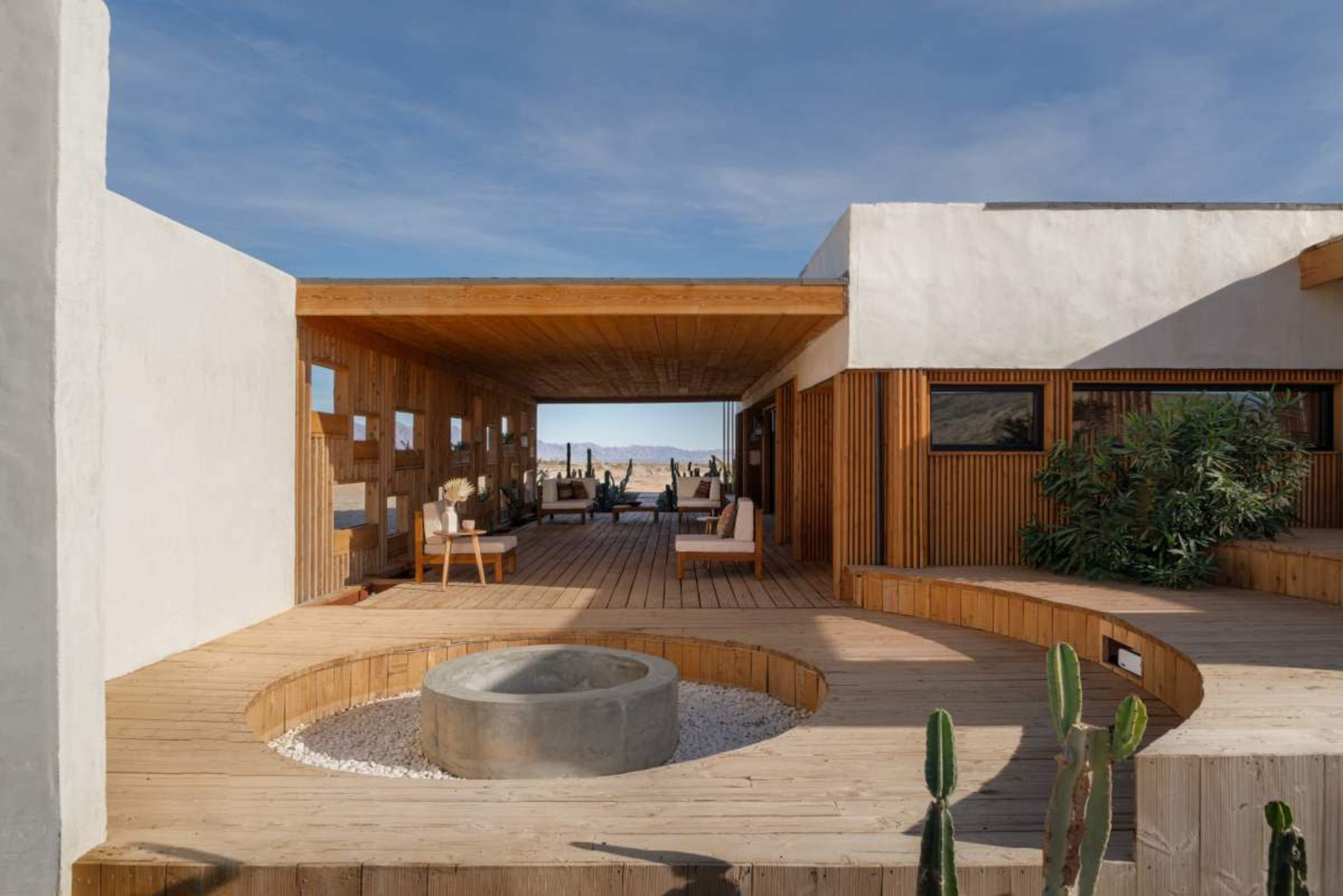 The image depicts a modern, single-story house with a wooden deck and an outdoor seating area, surrounded by minimalist landscaping and large windows that overlook a desert landscape.