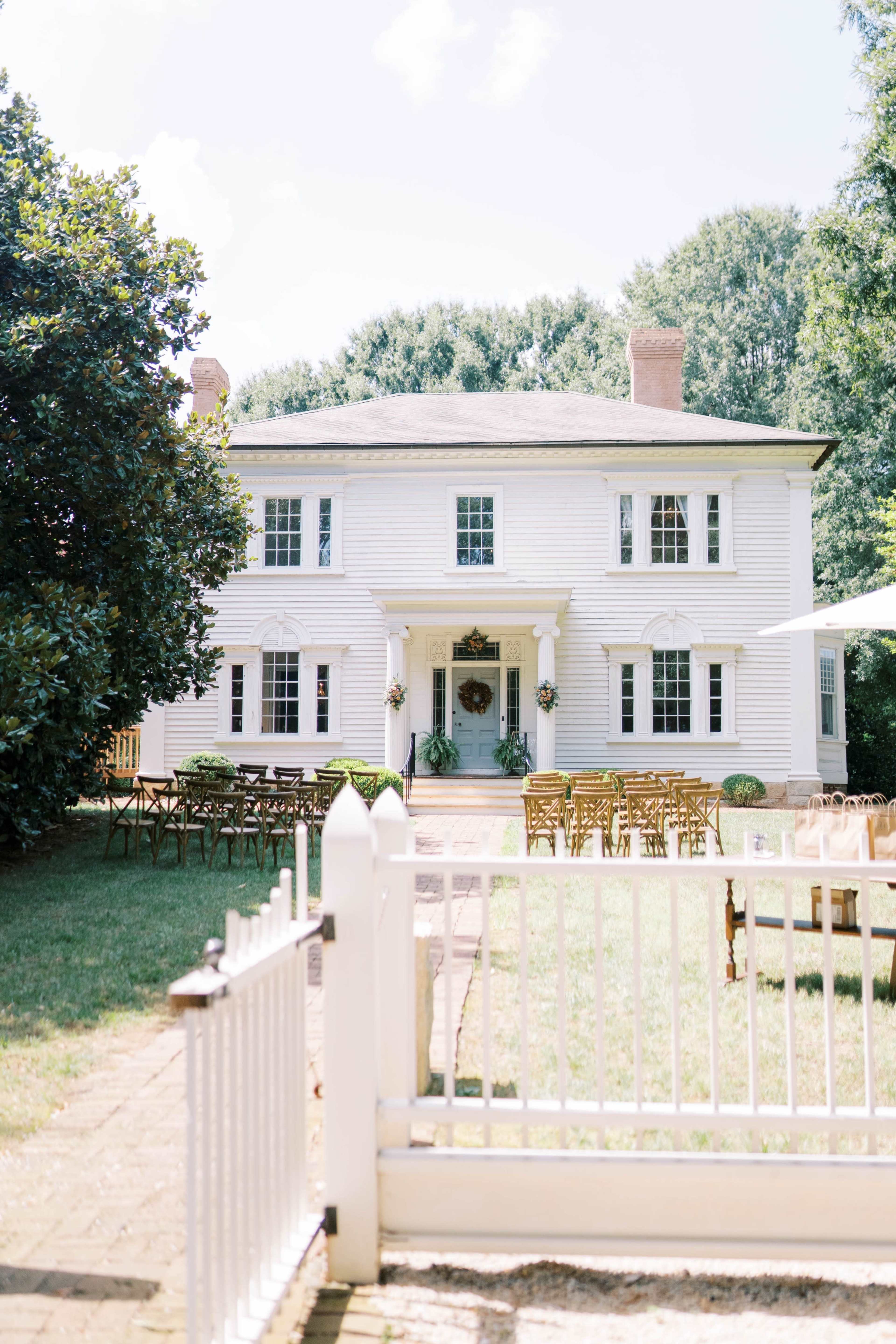 A white, two-story house with a central entrance is surrounded by chairs arranged for an event in the front yard.