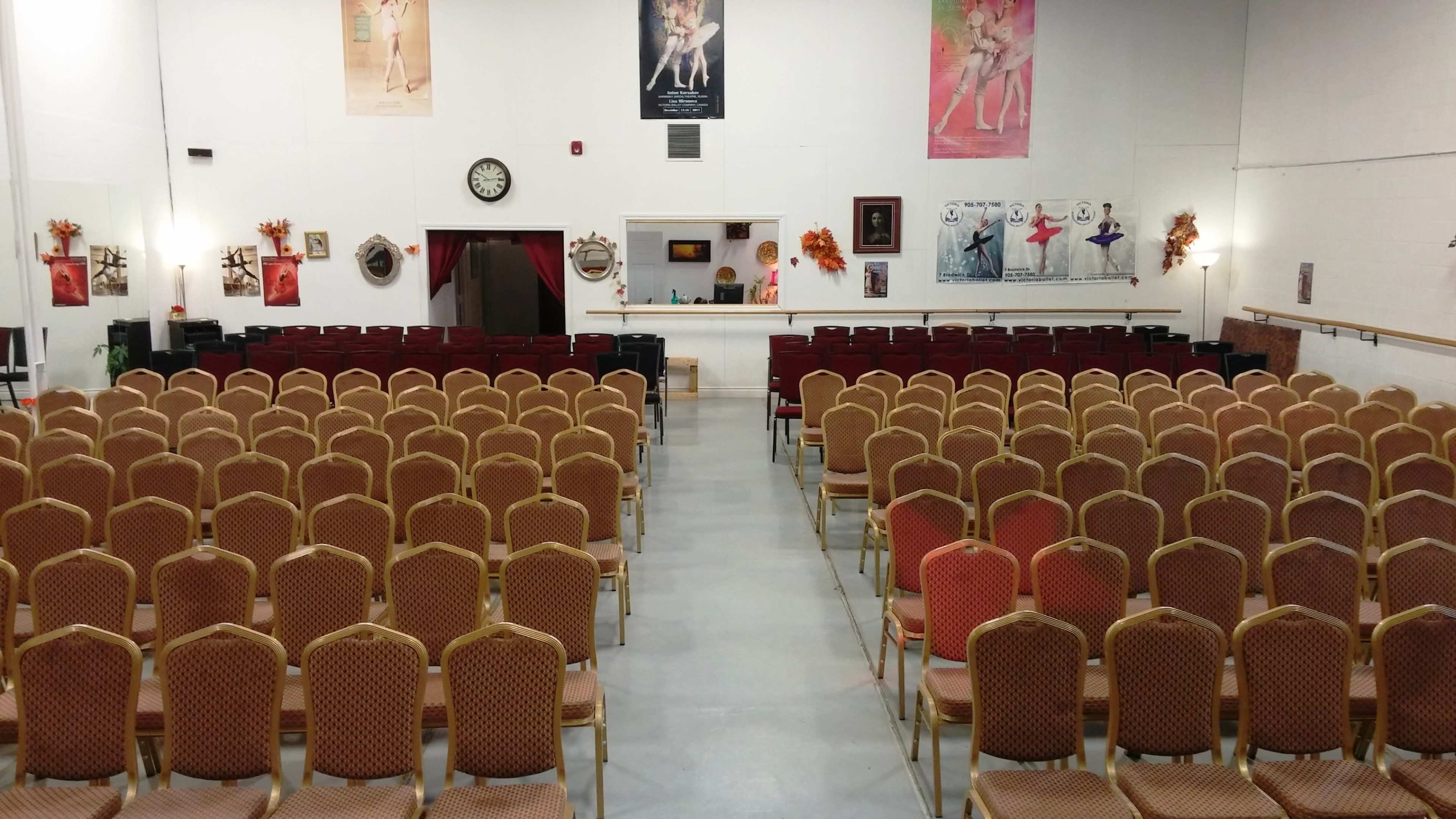 A large room with rows of empty, gold-framed chairs facing a stage area, decorated with various artworks and posters on the walls.