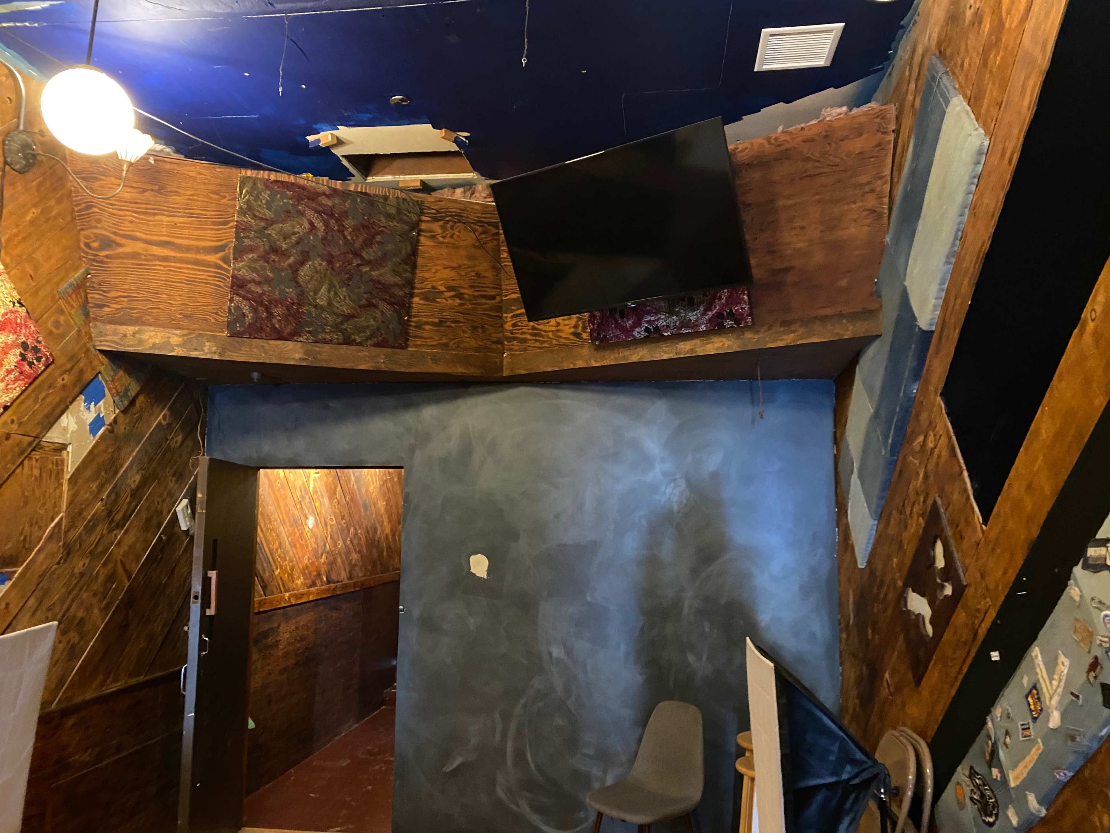 The interior of a room features a slanted wooden wall with patches of fabric and a mounted television, along with a blue chalkboard area and a chair in the corner.