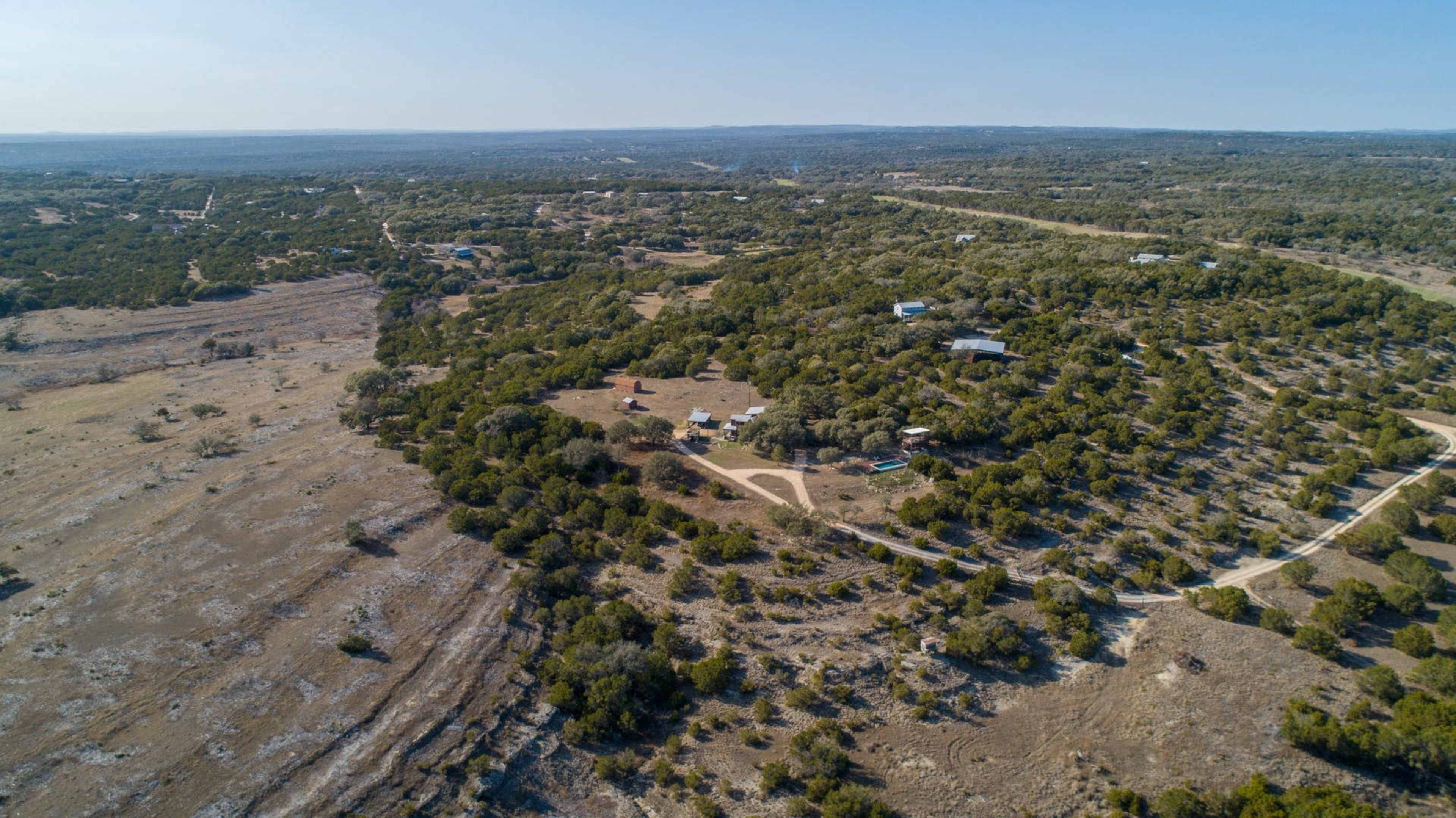 The image shows an aerial view of a hilly landscape with scattered trees and a few buildings amidst open land.