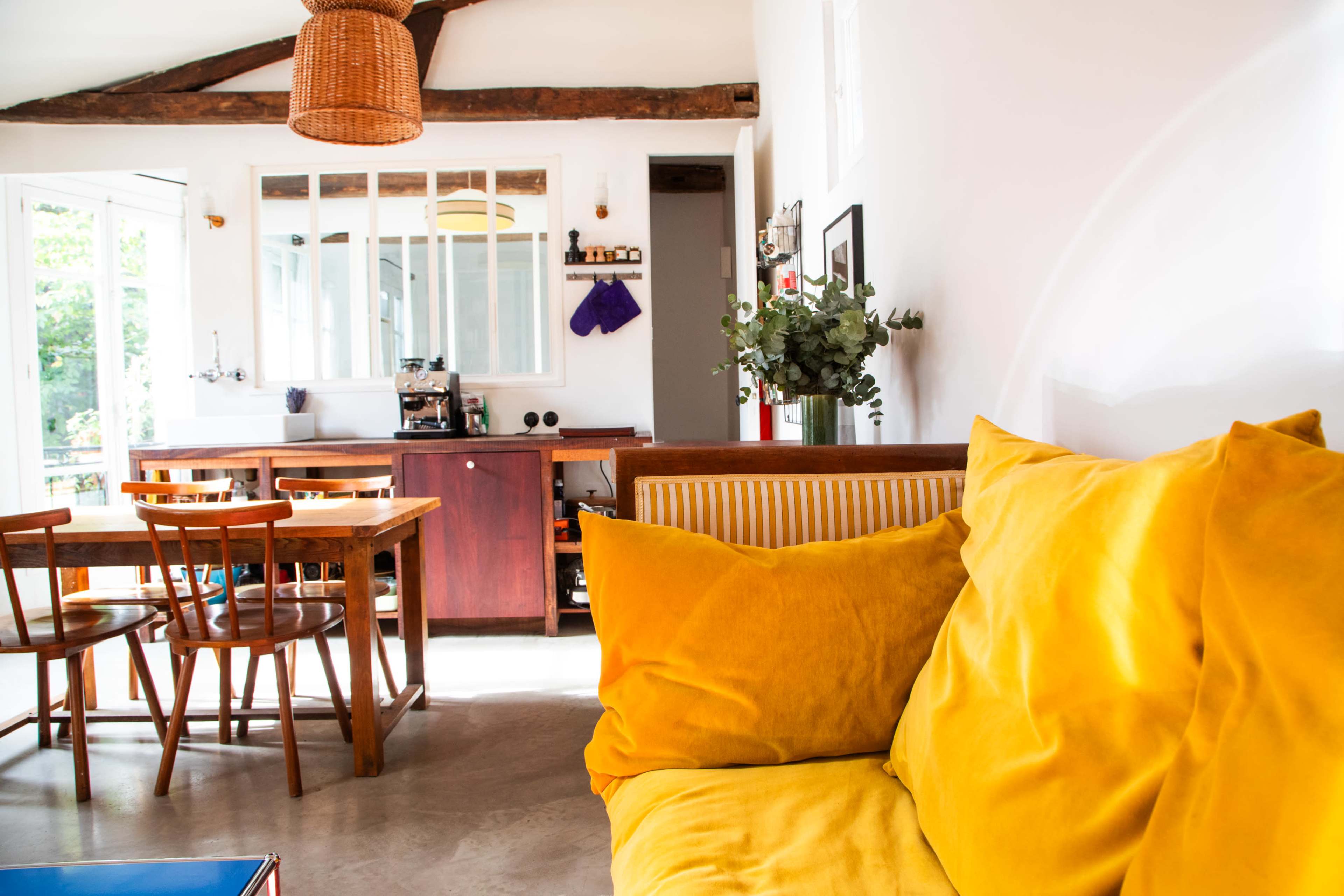 The image shows a bright, open living space with a yellow couch, a wooden dining table, and a kitchen area in the background.