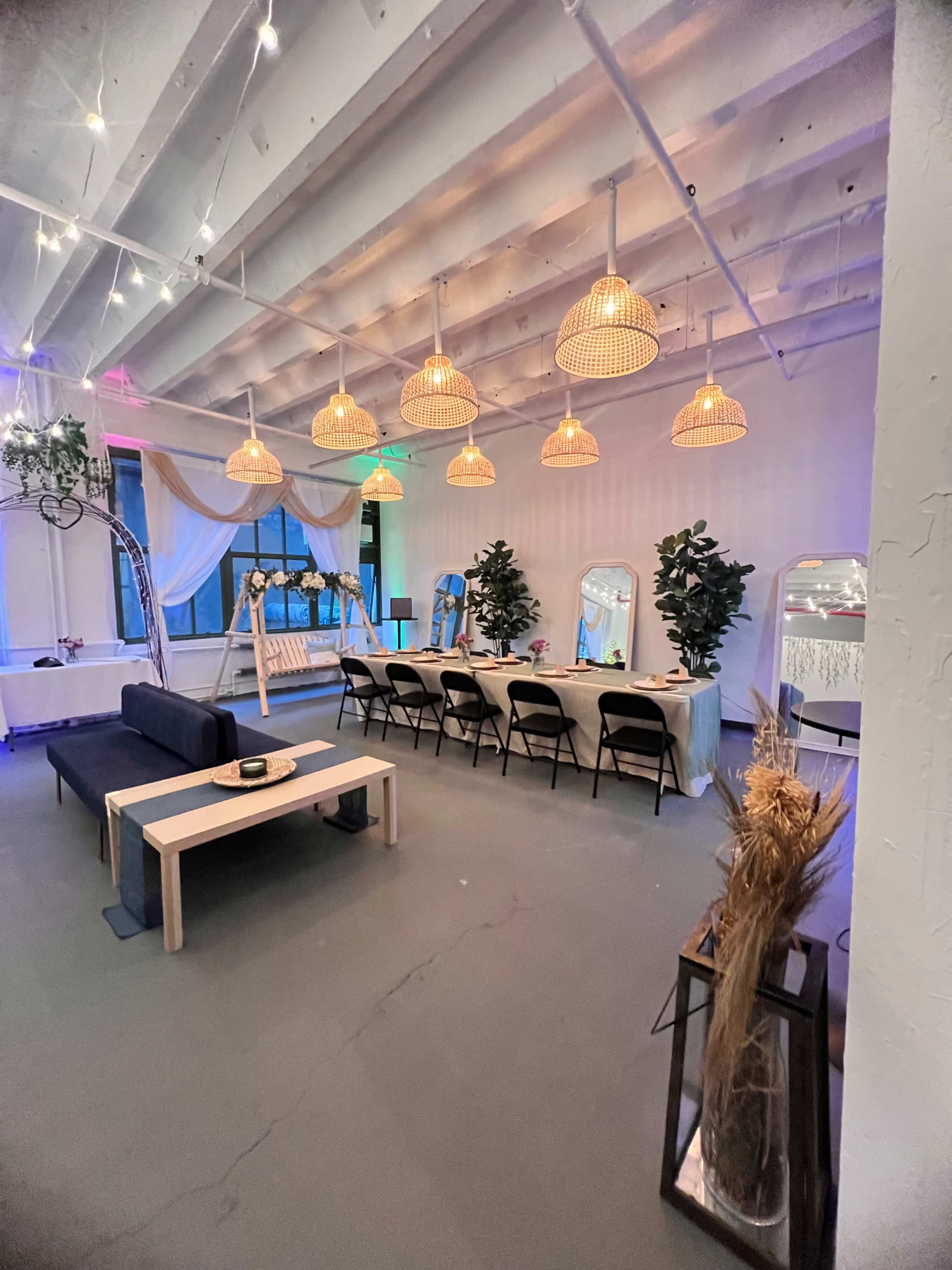 A modern event space with a long dining table set for guests, surrounded by potted plants and decorated with hanging wicker lights.