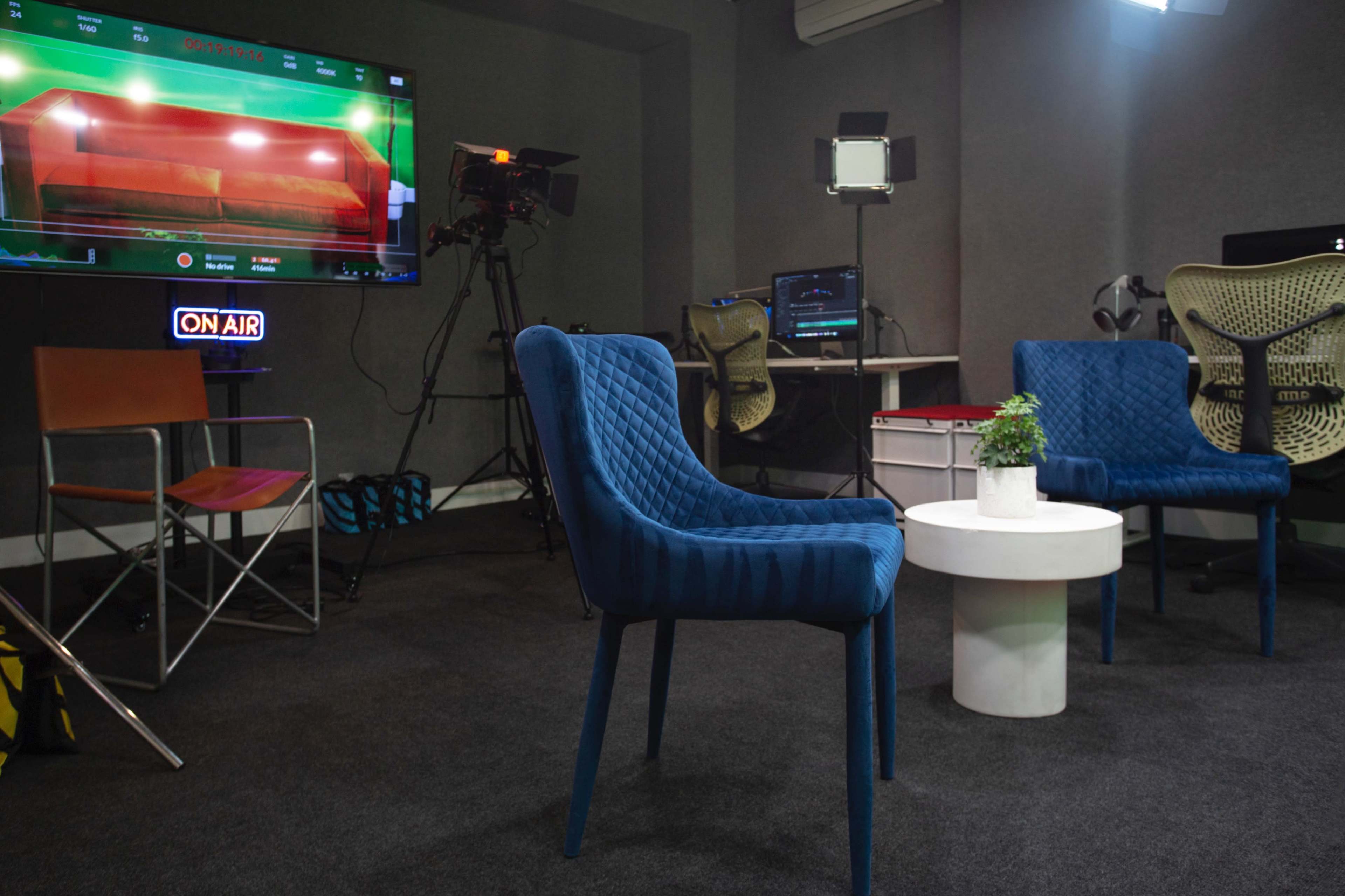 A media studio setup featuring two blue chairs, a small white table, and various broadcasting equipment in the background.