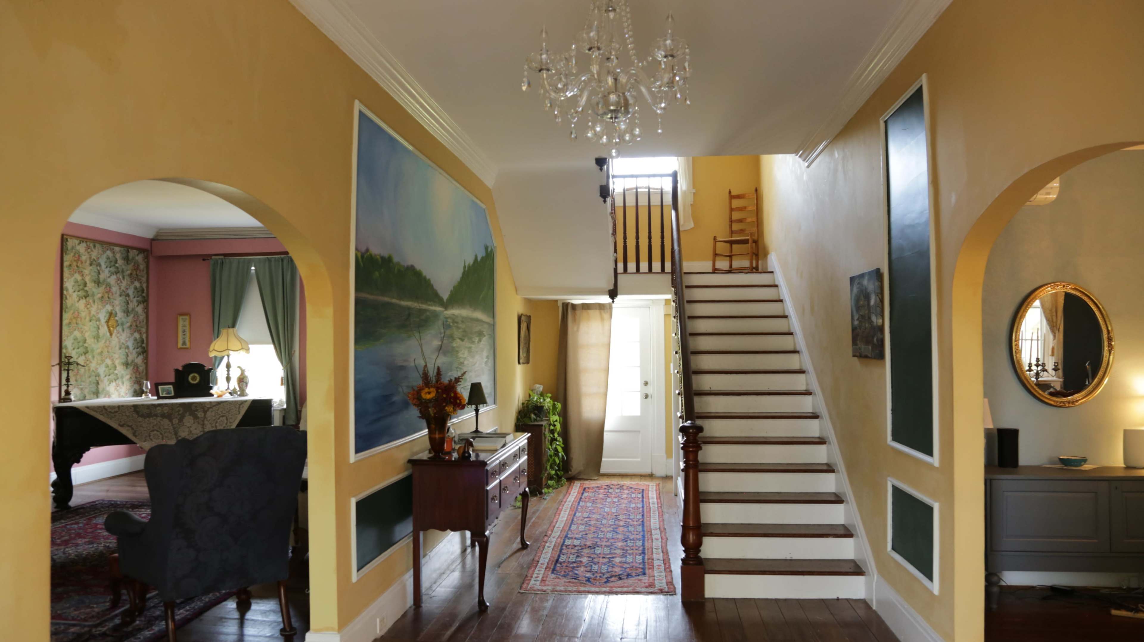 A brightly lit foyer with a staircase leading to an upper level, flanked by a large painting and decorative furnishings.