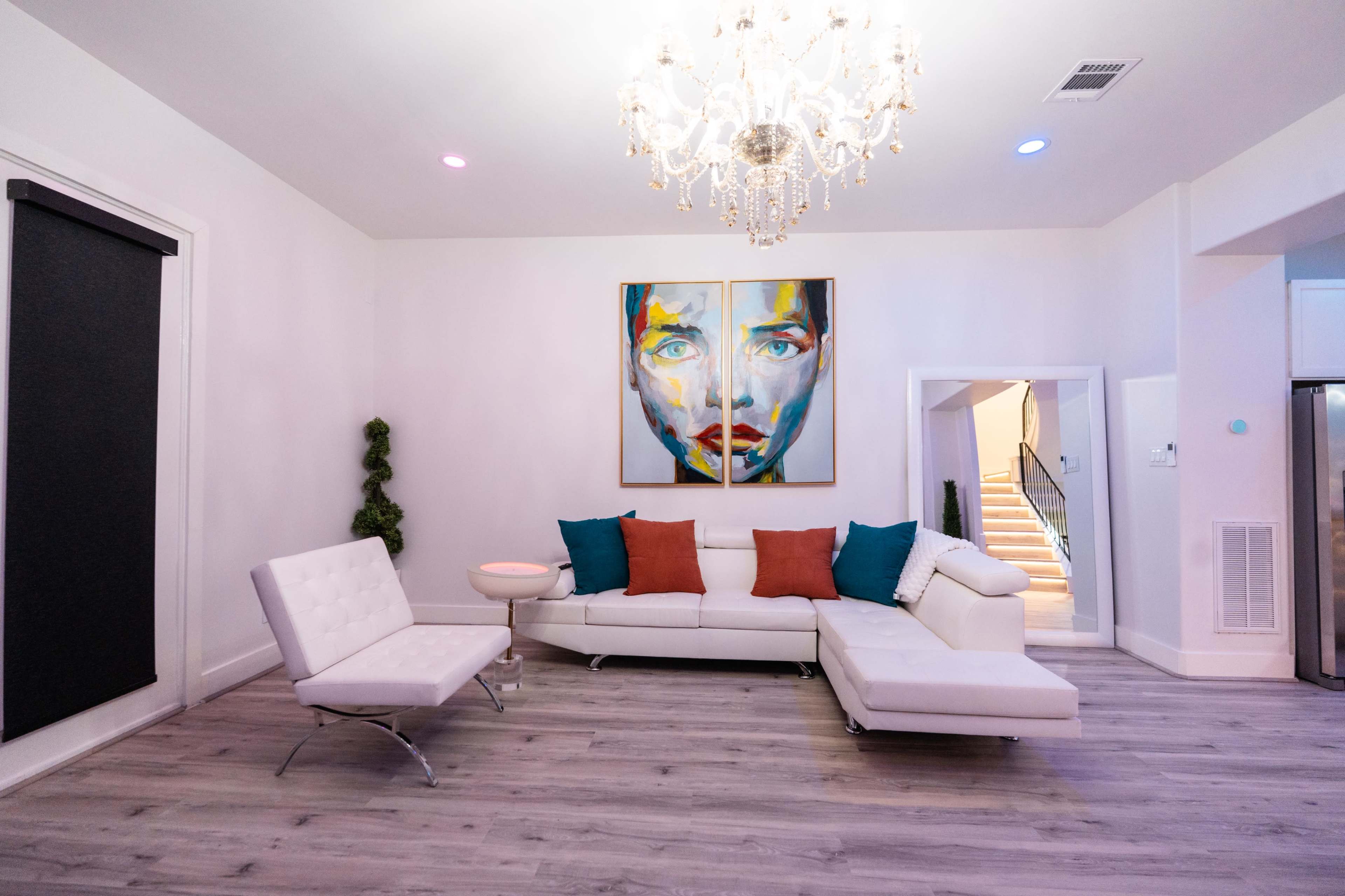 The image shows a modern living room featuring a white sectional sofa, a large chandelier, and vibrant artwork on the walls.
