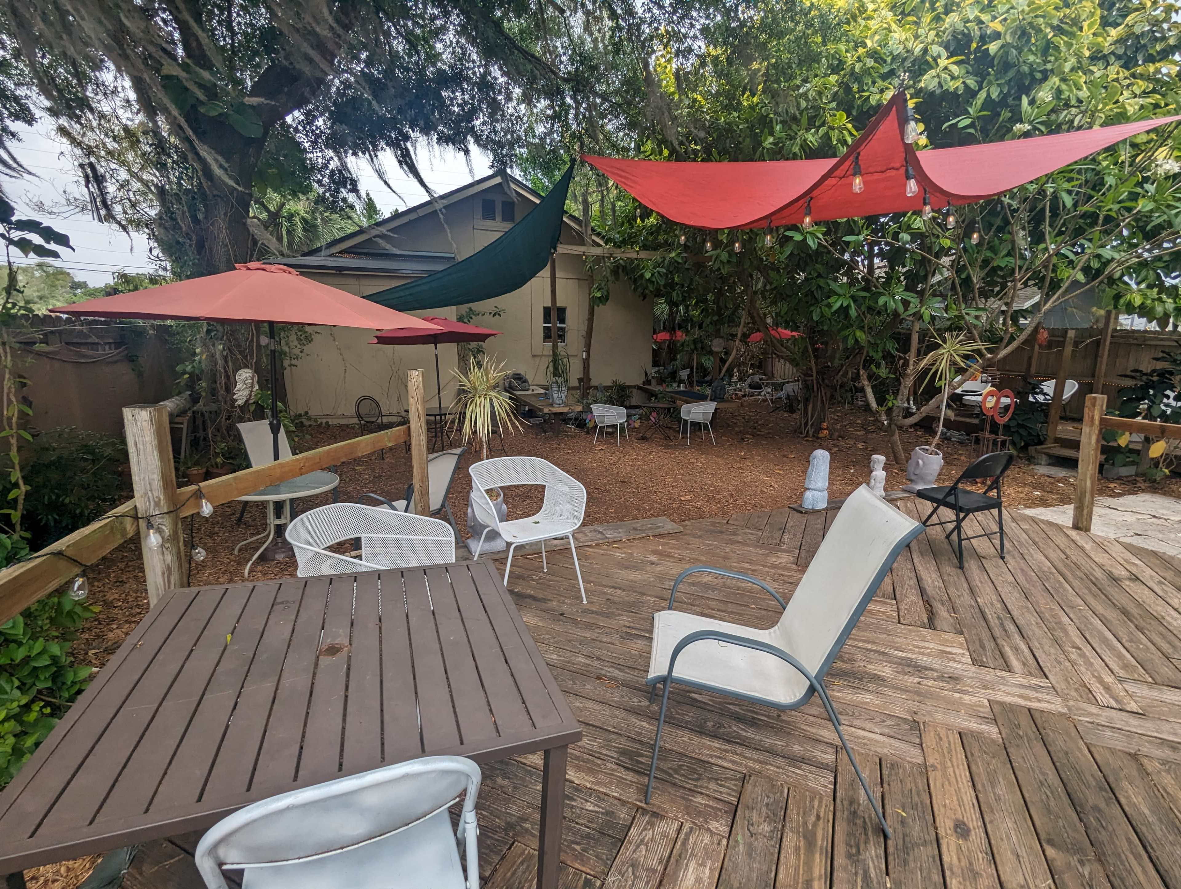 The image shows a serene outdoor seating area with wooden decking, various chairs, and colorful fabric shades overhead amid lush greenery.
