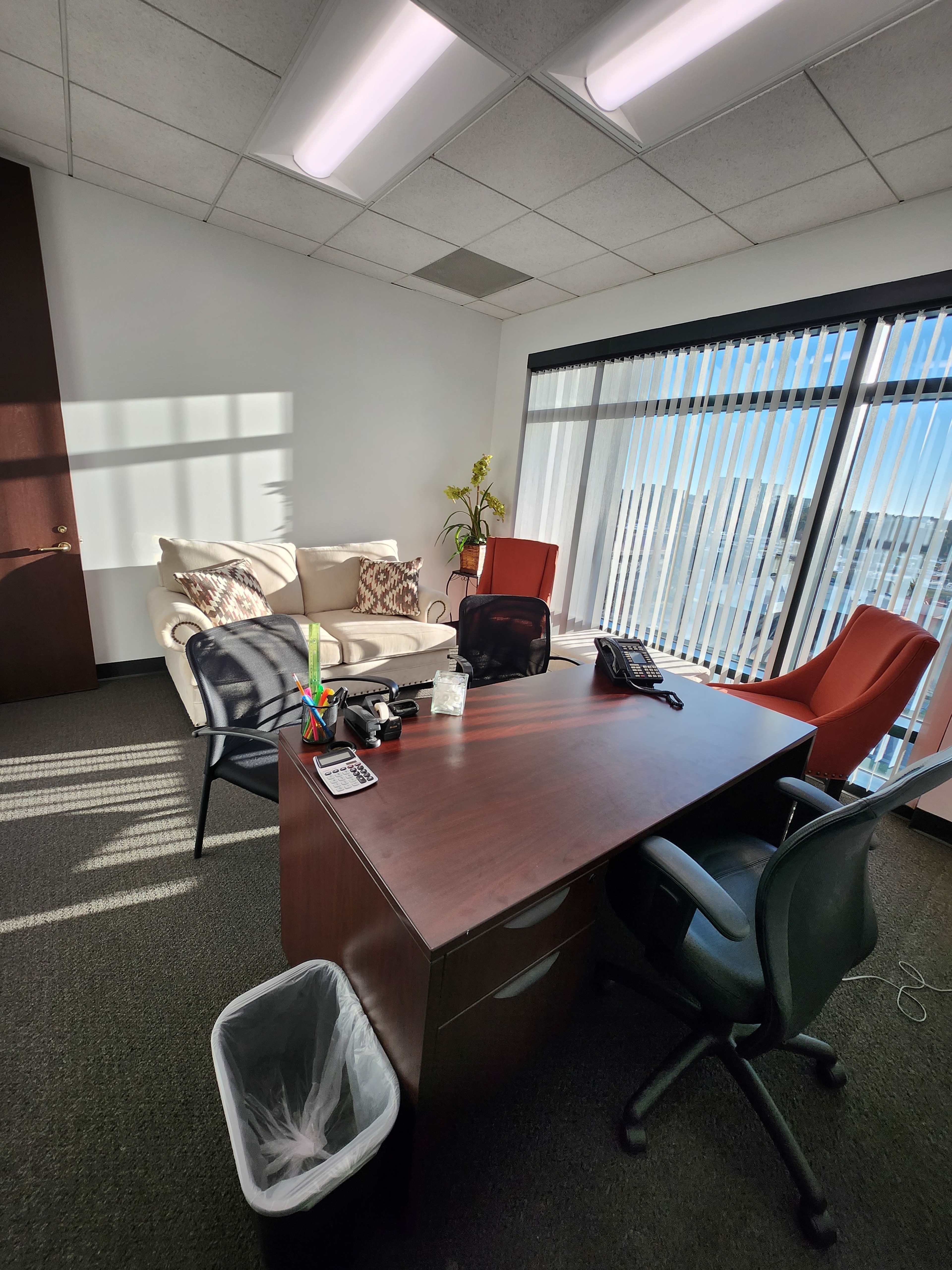The image shows an office space with a wooden desk, two office chairs, a wastebasket, a sofa, and large windows with vertical blinds allowing natural light to enter.