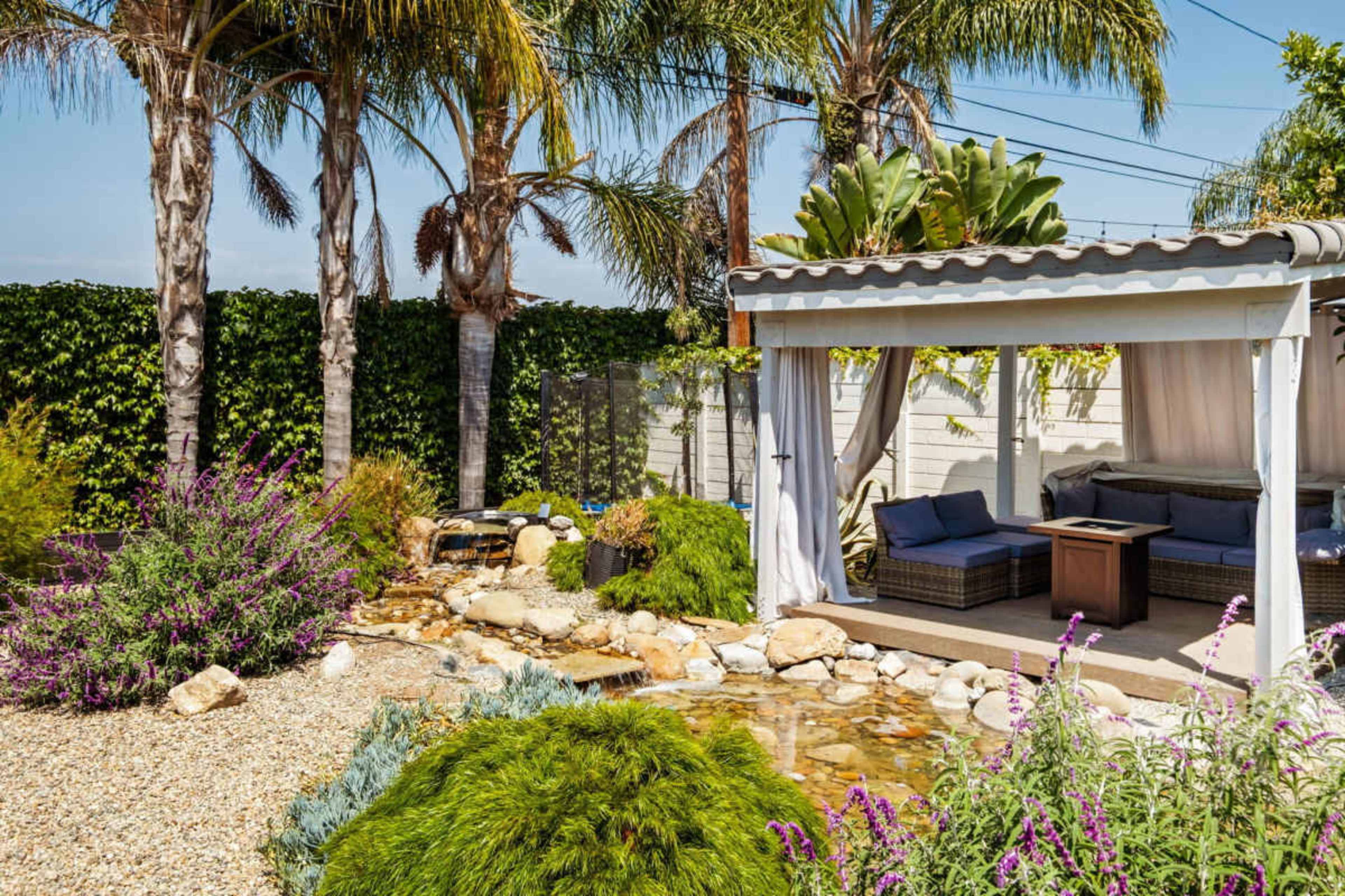 A landscaped garden features a gazebo with outdoor seating beside a small stream and lush greenery, including palm trees and colorful flowers.