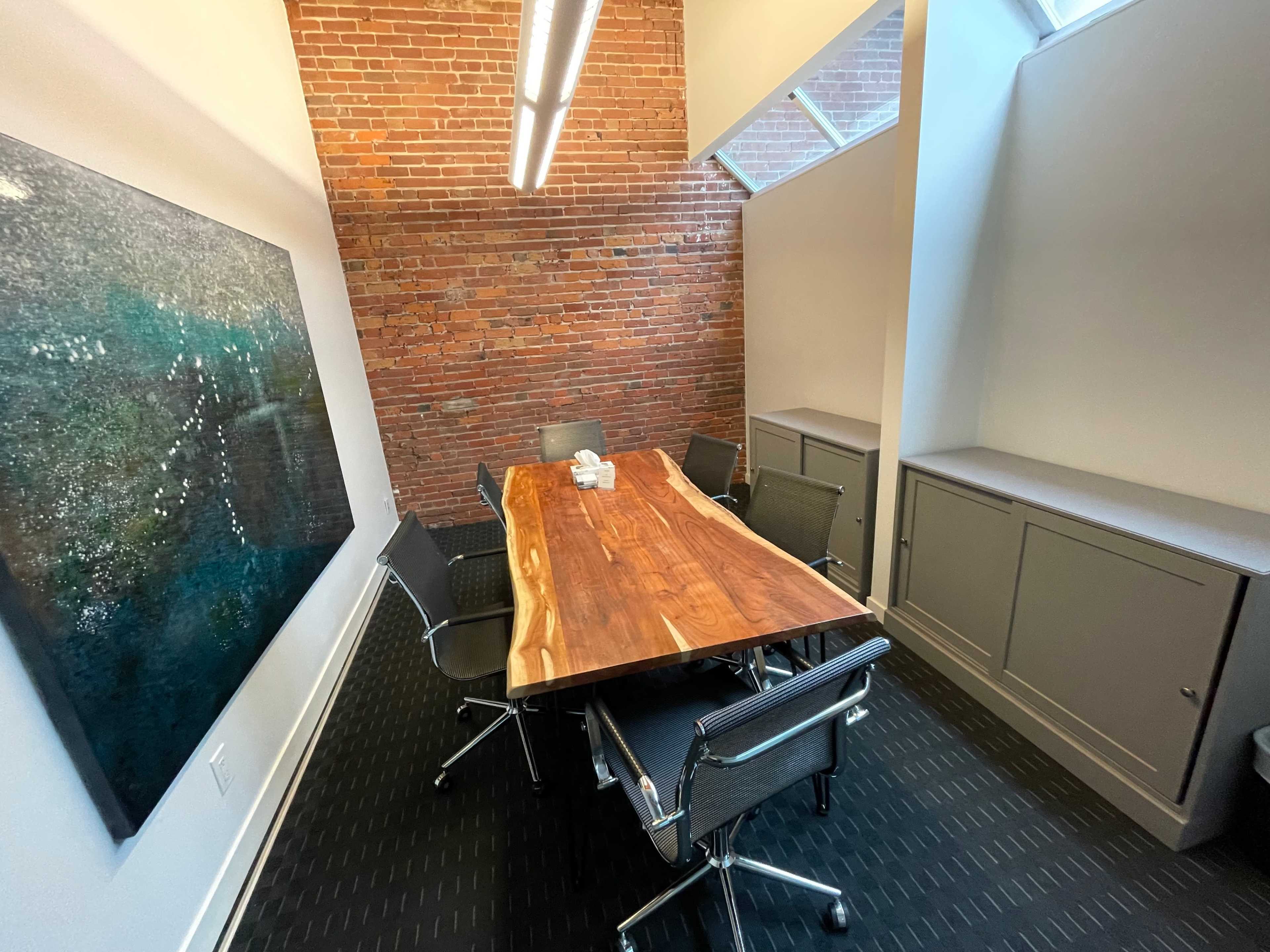 A conference room features a wooden table surrounded by six metal chairs, with a large artwork on the wall and a brick accent wall.