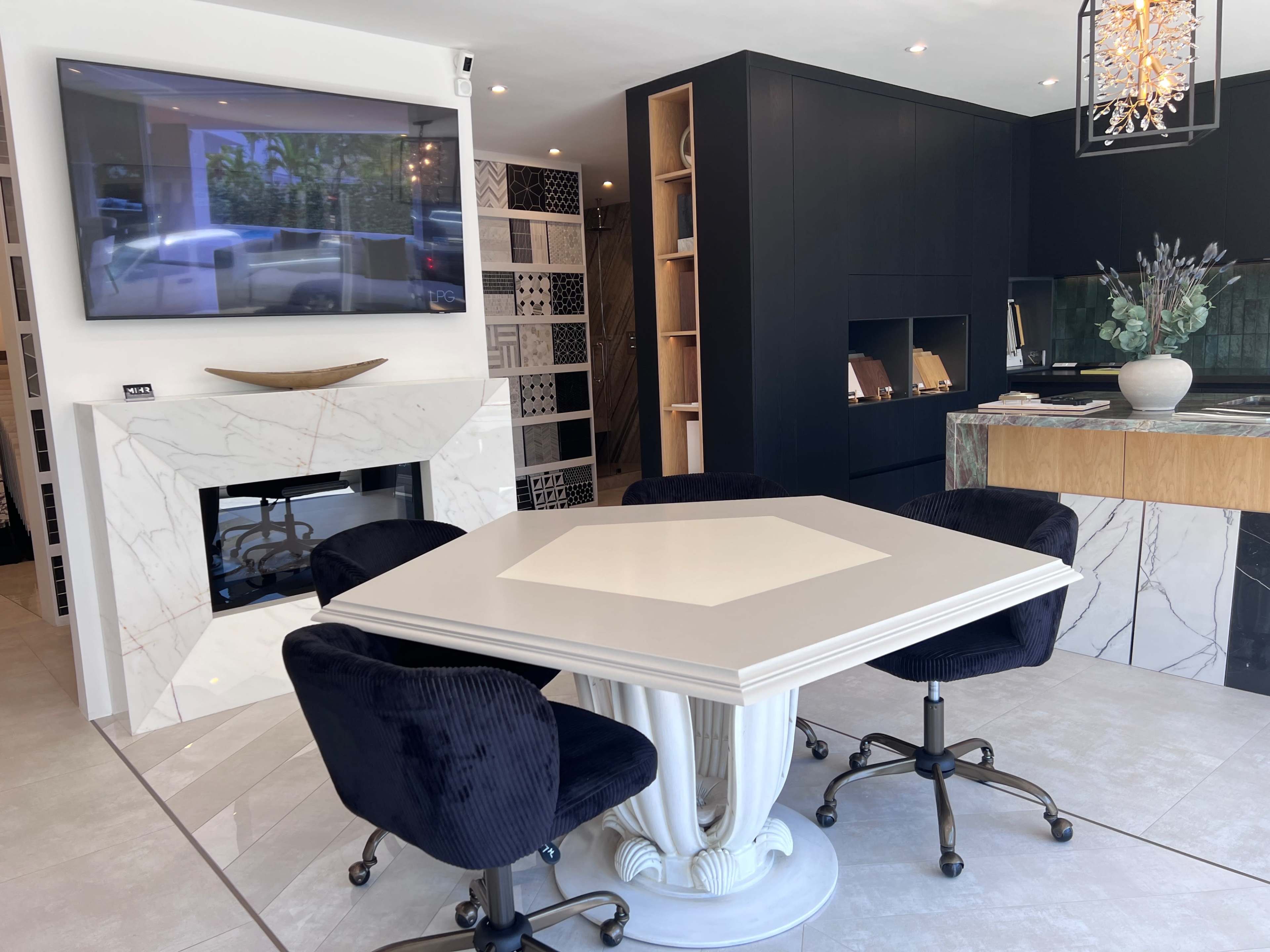 The image shows a modern interior space featuring a square table surrounded by black upholstered chairs, a television mounted on the wall, and a fireplace set into a marble wall.