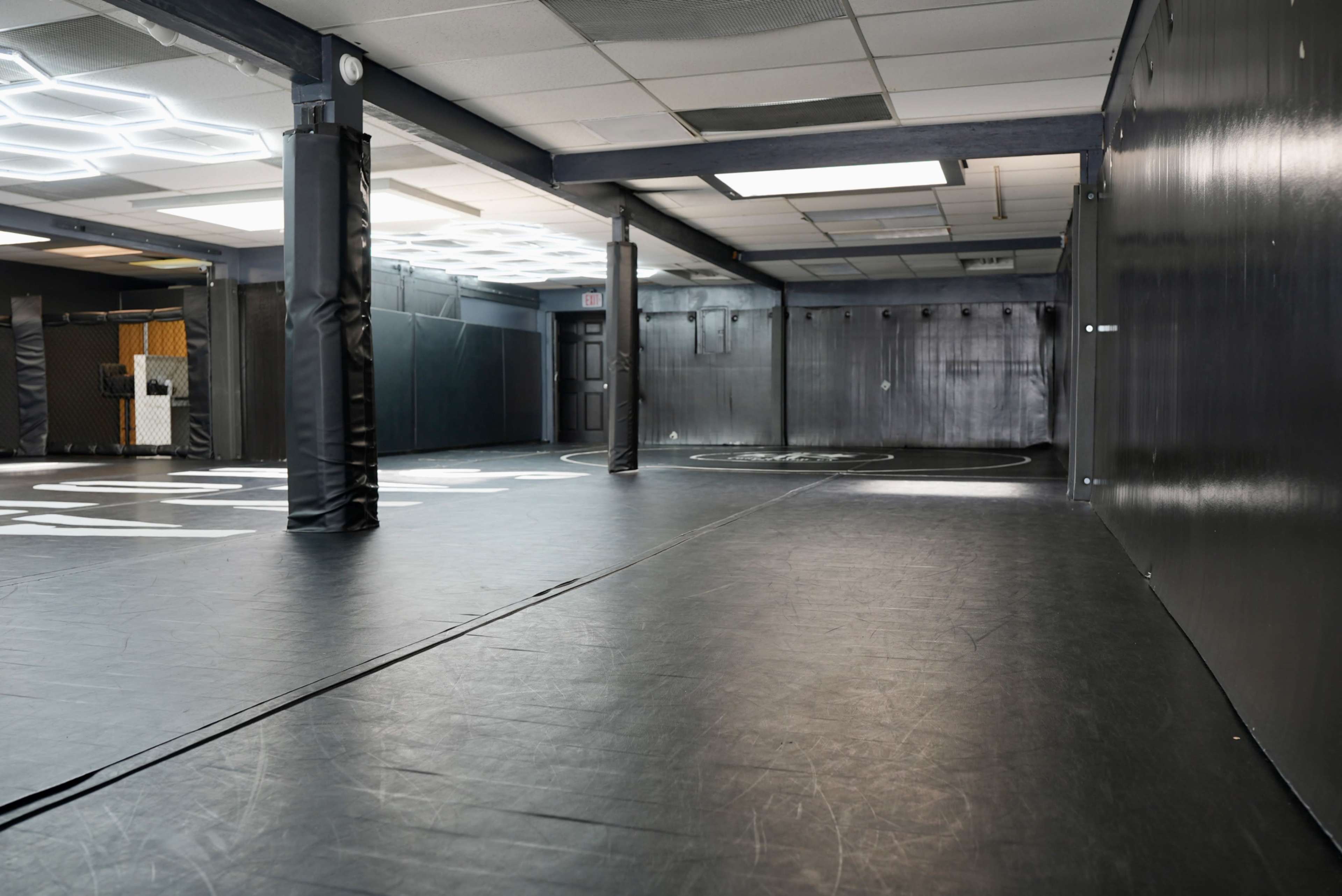 The image shows an empty training facility with a padded black floor and several cylindrical posts, illuminated by overhead lighting.