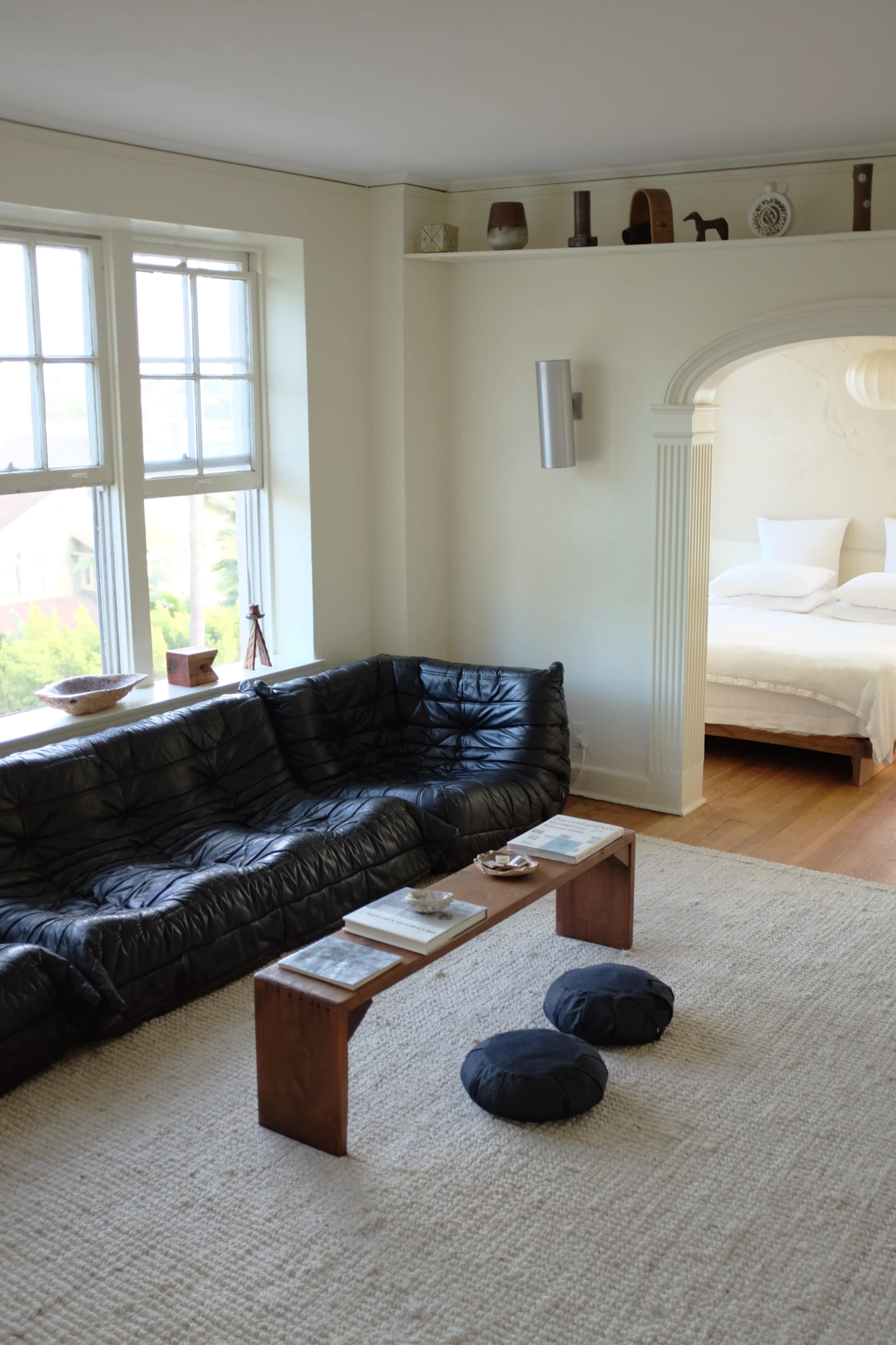 The image shows a cozy living room with a black leather sofa, a wooden coffee table, and cushioned stools, adjacent to an archway leading to a bedroom.