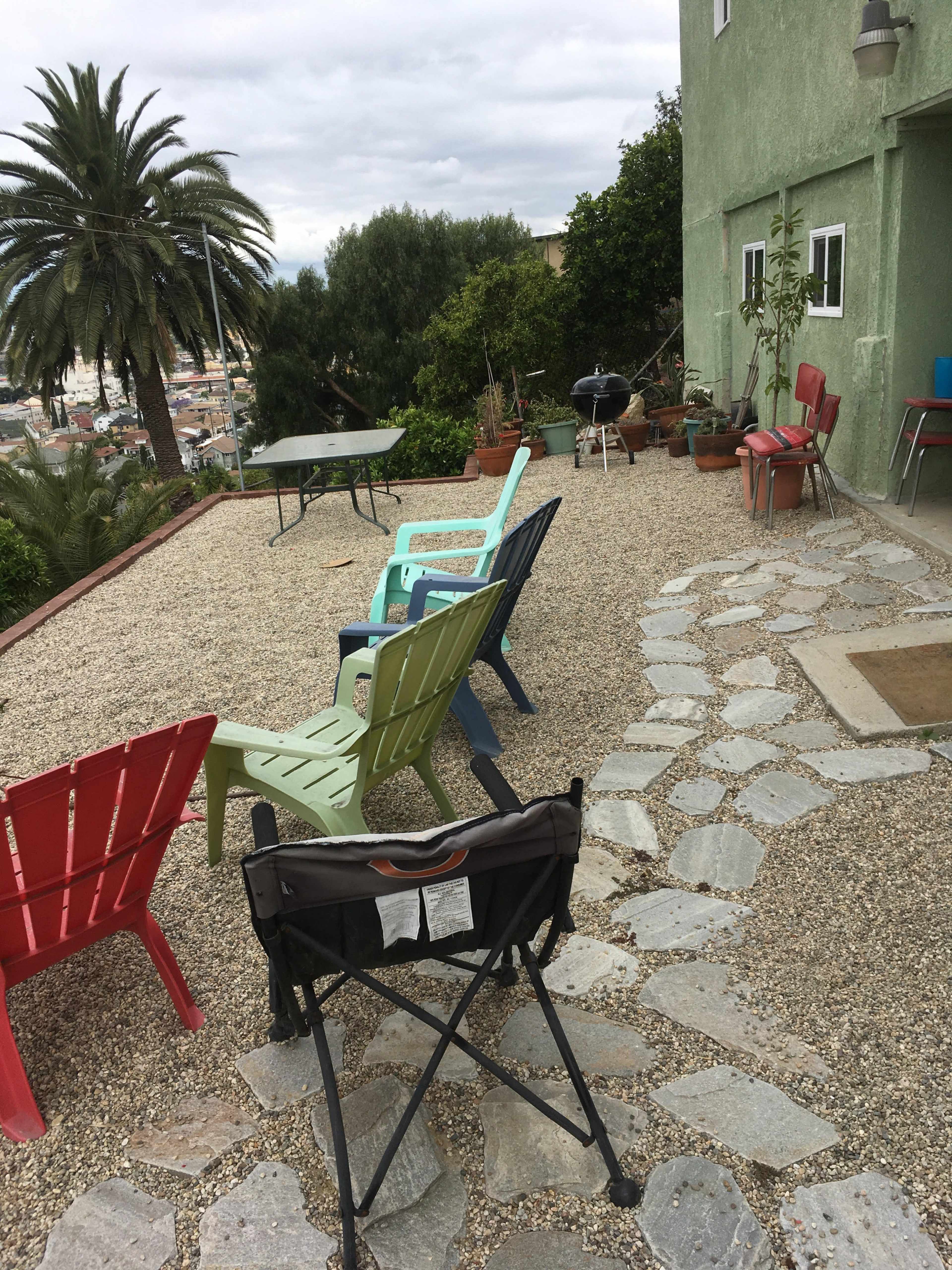 A patio area with chairs in various colors arranged near a gravel pathway, leading to a house surrounded by green plants and palm trees.