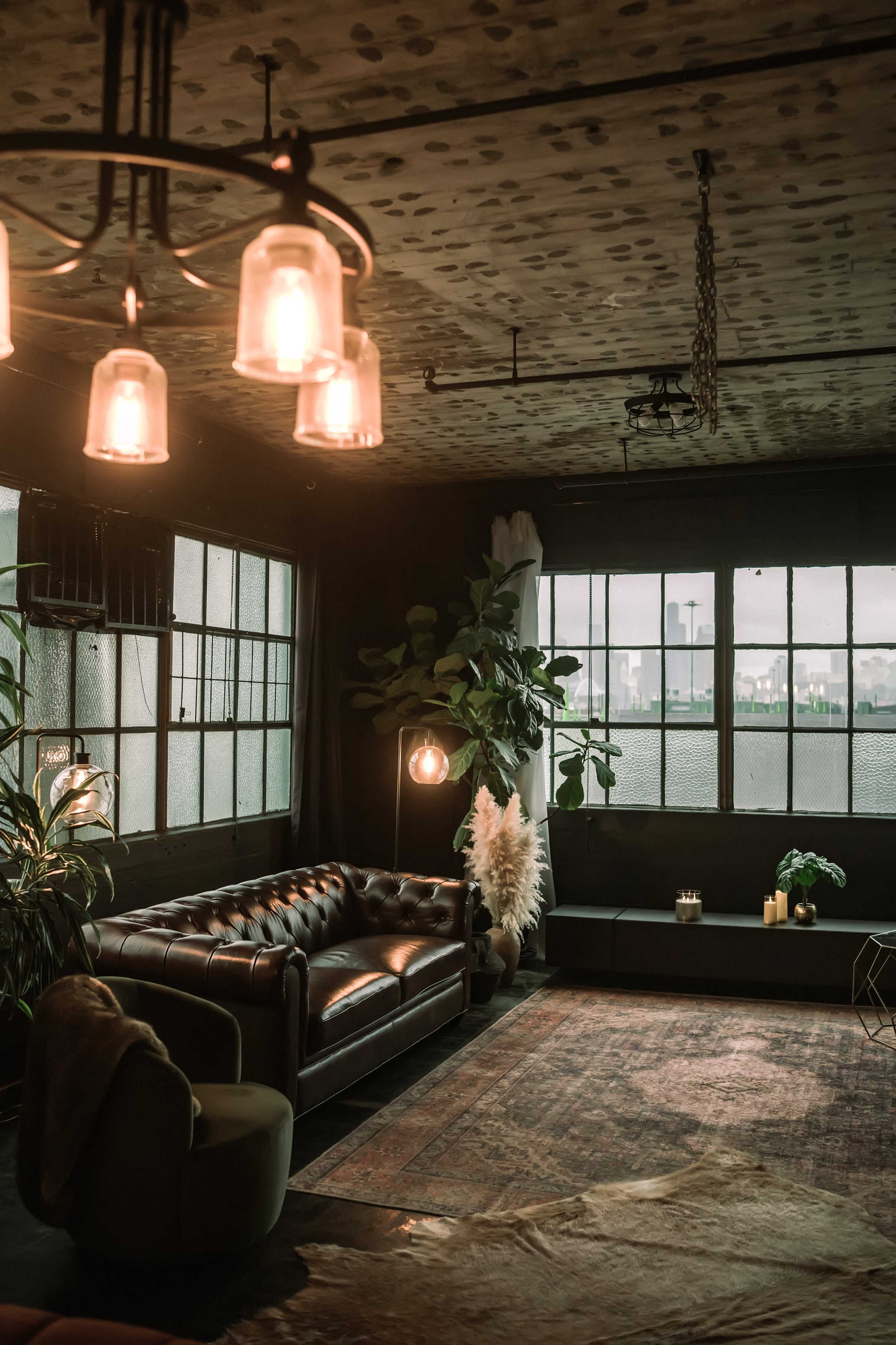 The image features a dimly lit interior with a leather sofa, large windows offering a view of a city skyline, and various plants and decor items.