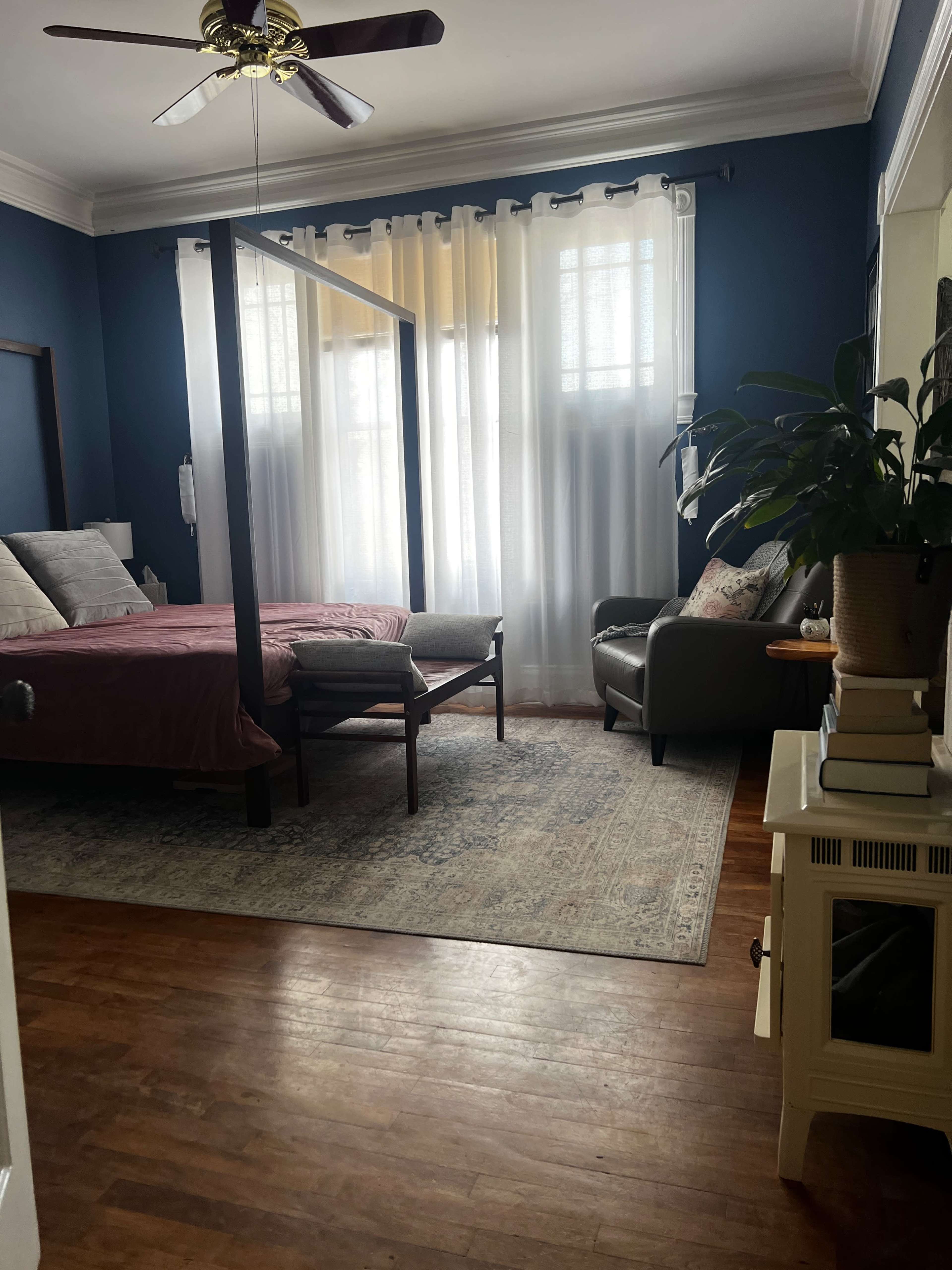 The image shows a bedroom with a four-poster bed, a seating area with an armchair, and large windows dressed in sheer curtains, creating a bright and open space.