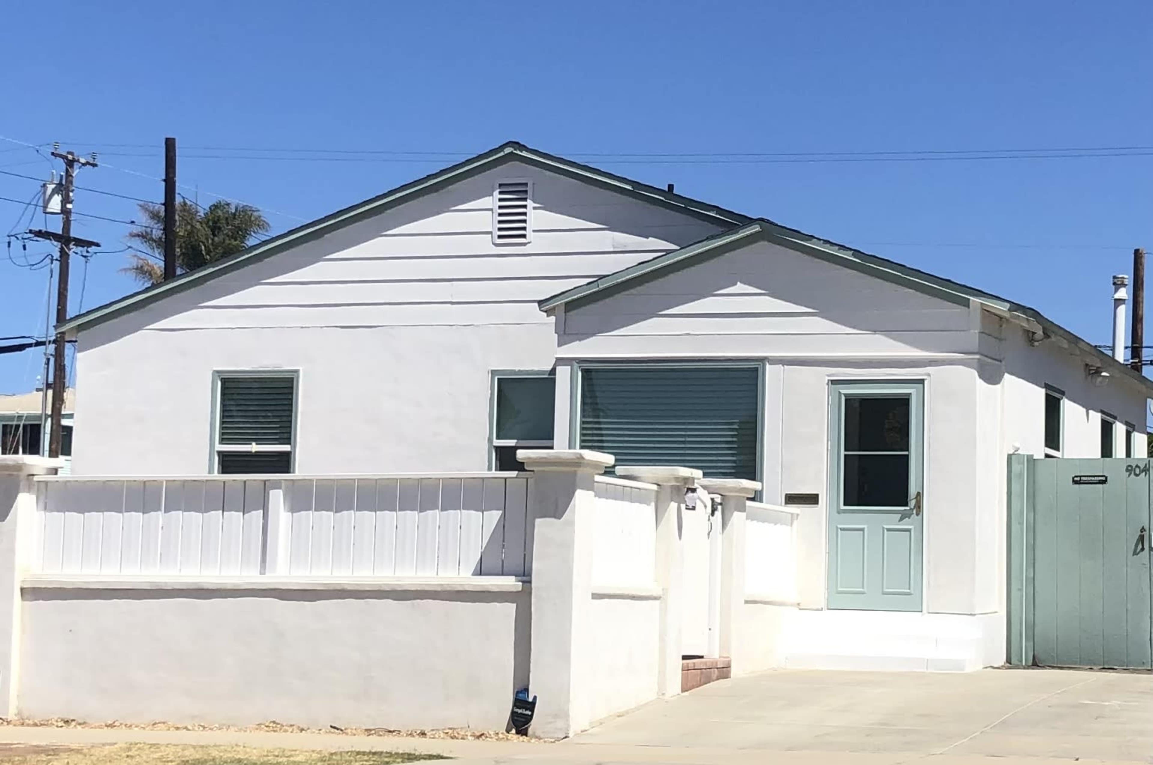 The image shows a white, single-story house with green trim and closed shutters, situated on a corner lot with a fence.