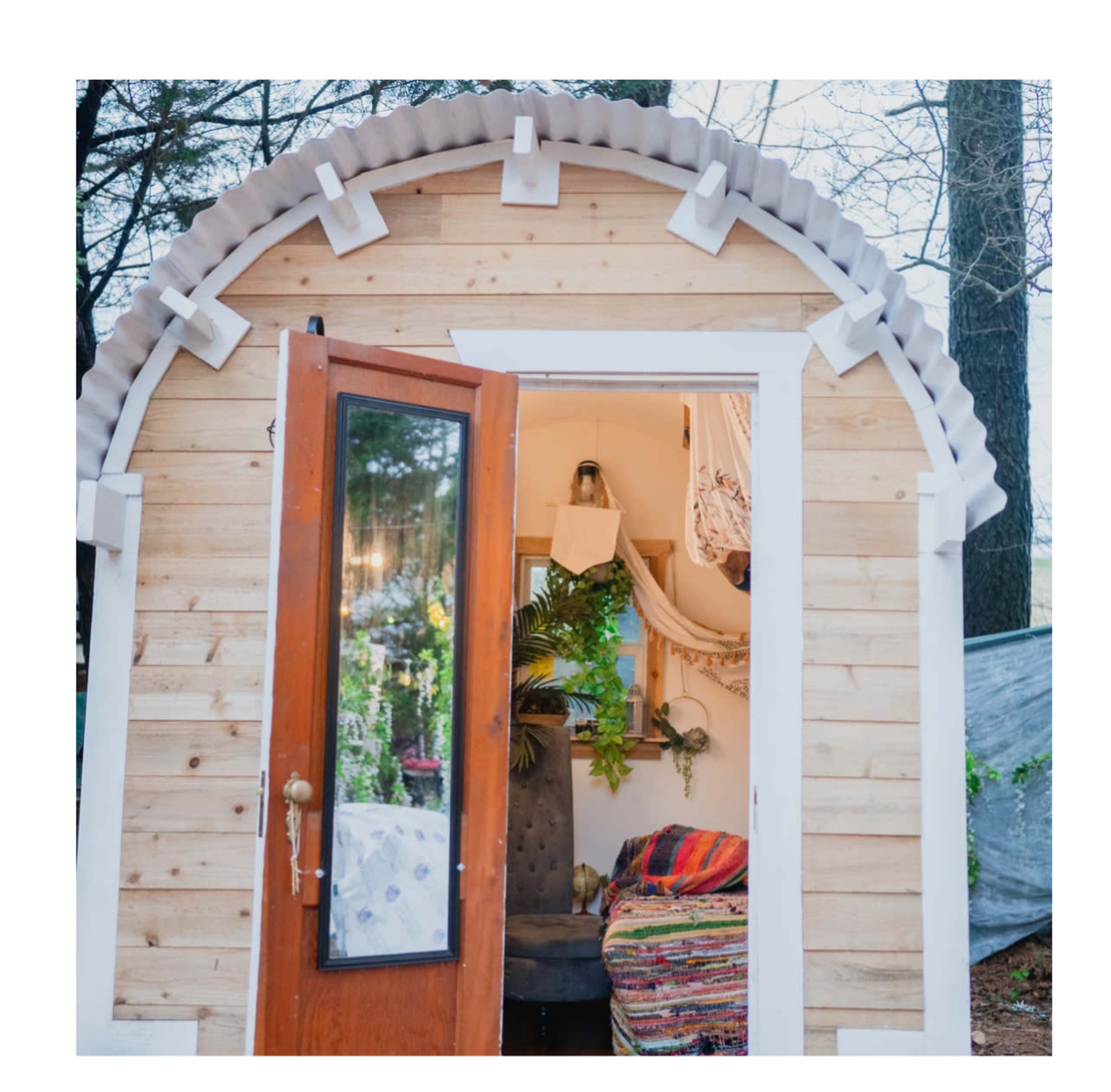 A small wooden cabin with a curved roof and open door, revealing a colorful interior decorated with plants and textiles.