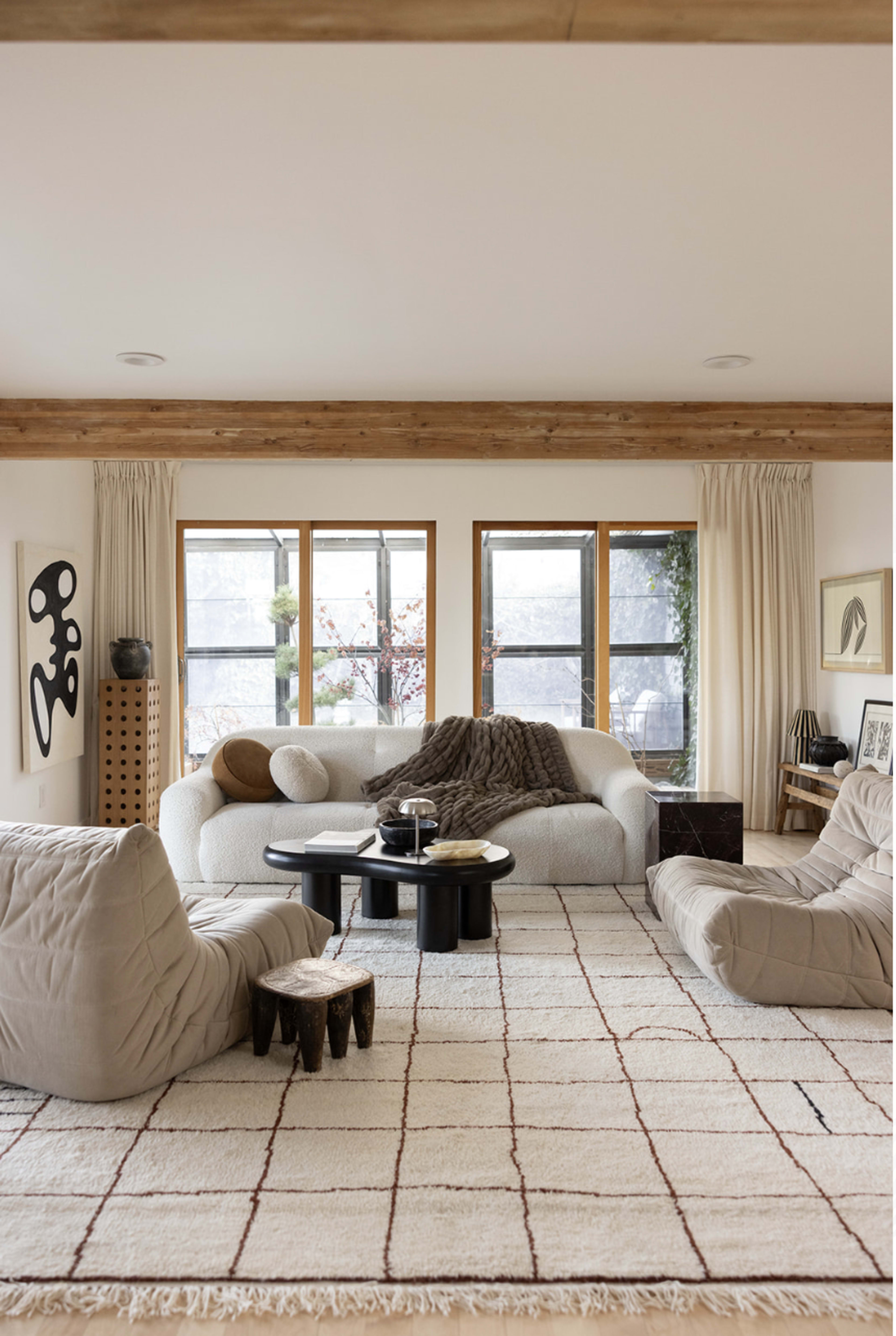 The living room features a light-colored sectional sofa, two oversized chairs, and a round black coffee table, all set on a patterned rug with large windows letting in natural light.