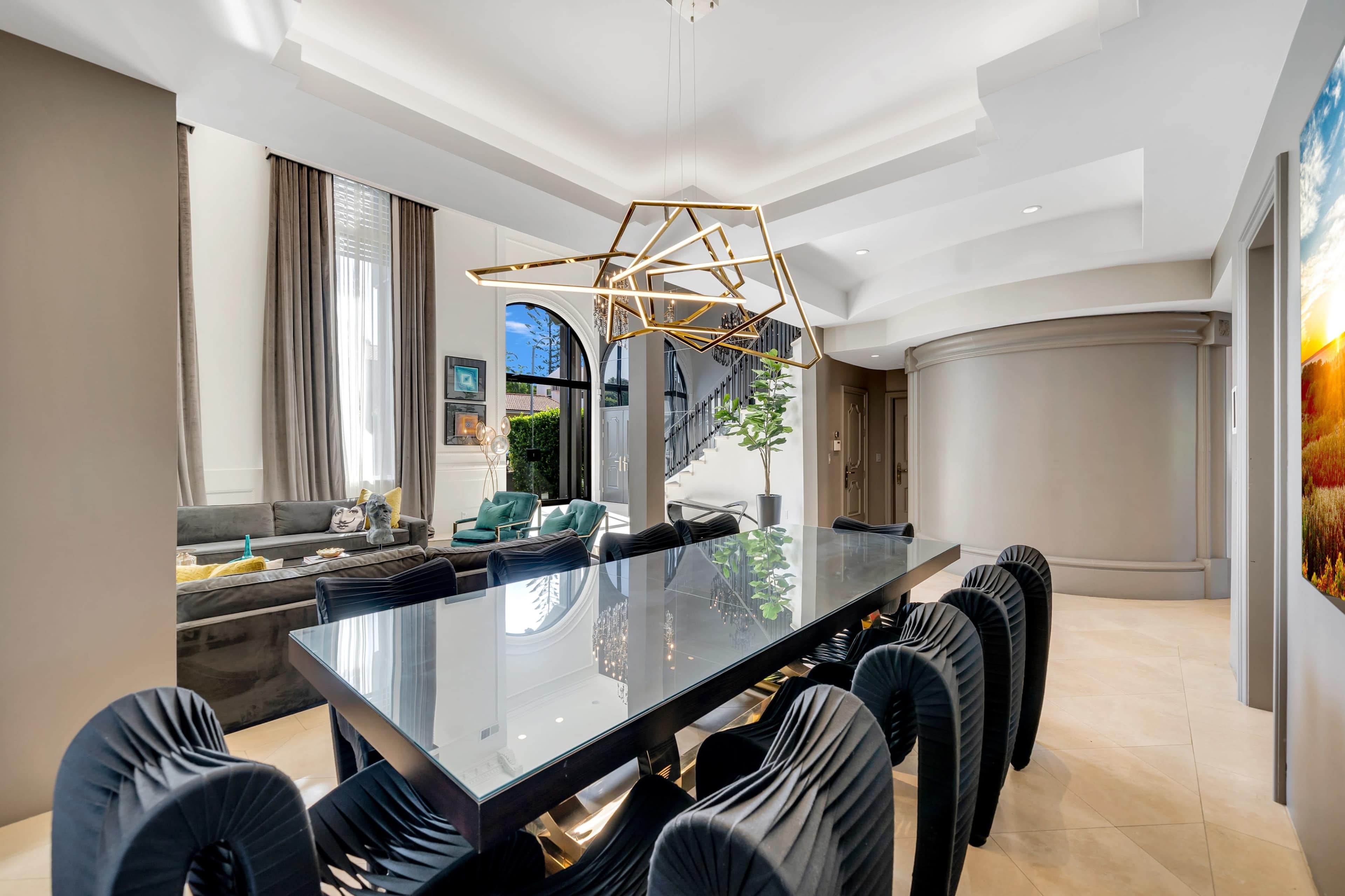 A modern dining area with a large glass table surrounded by black chairs, illuminated by a unique geometric chandelier, and leads to a staircase in a well-lit space.