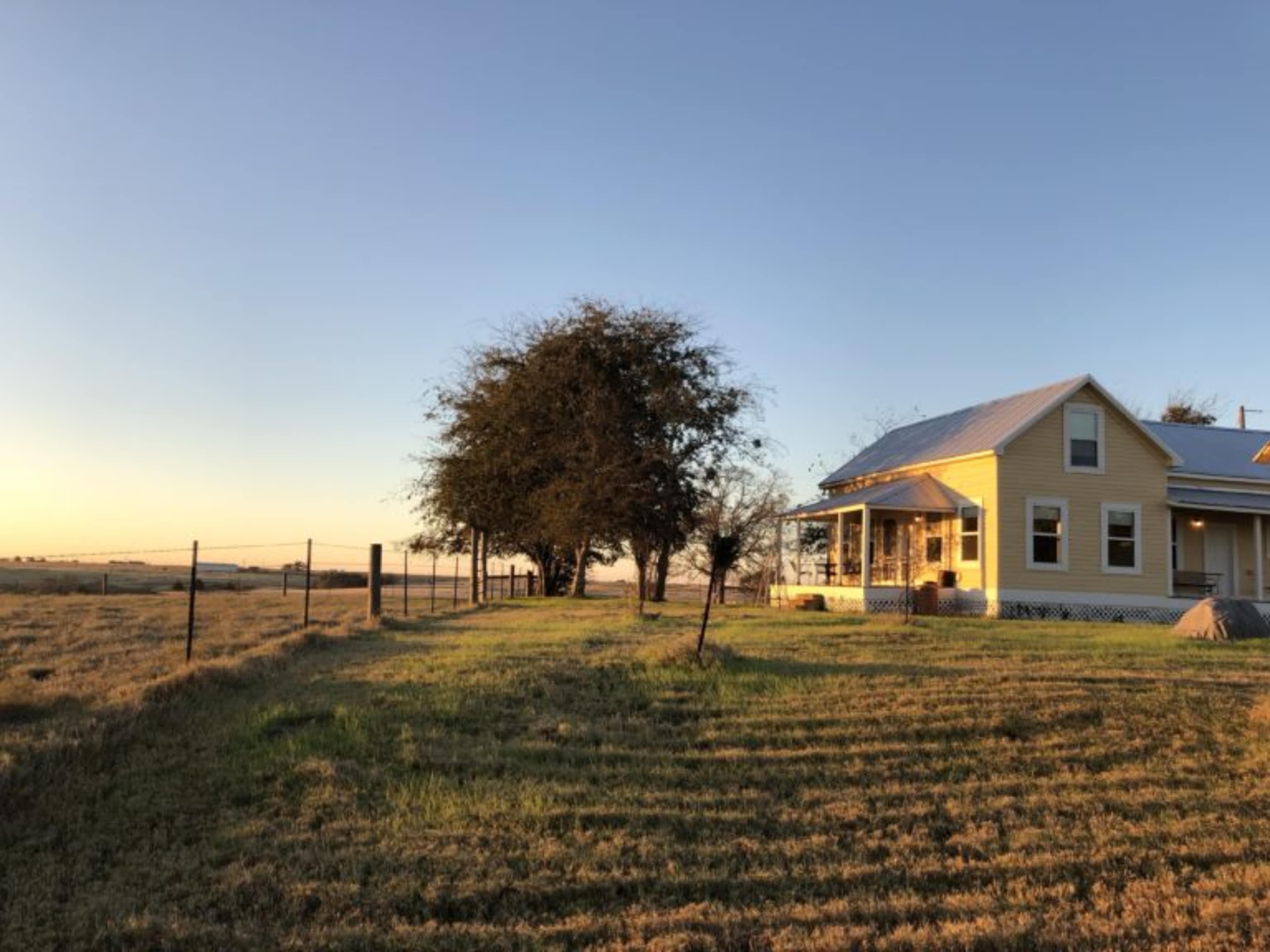 A yellow farmhouse is situated on a grassy hill, with a fence and a few trees in the background under a clear sky.