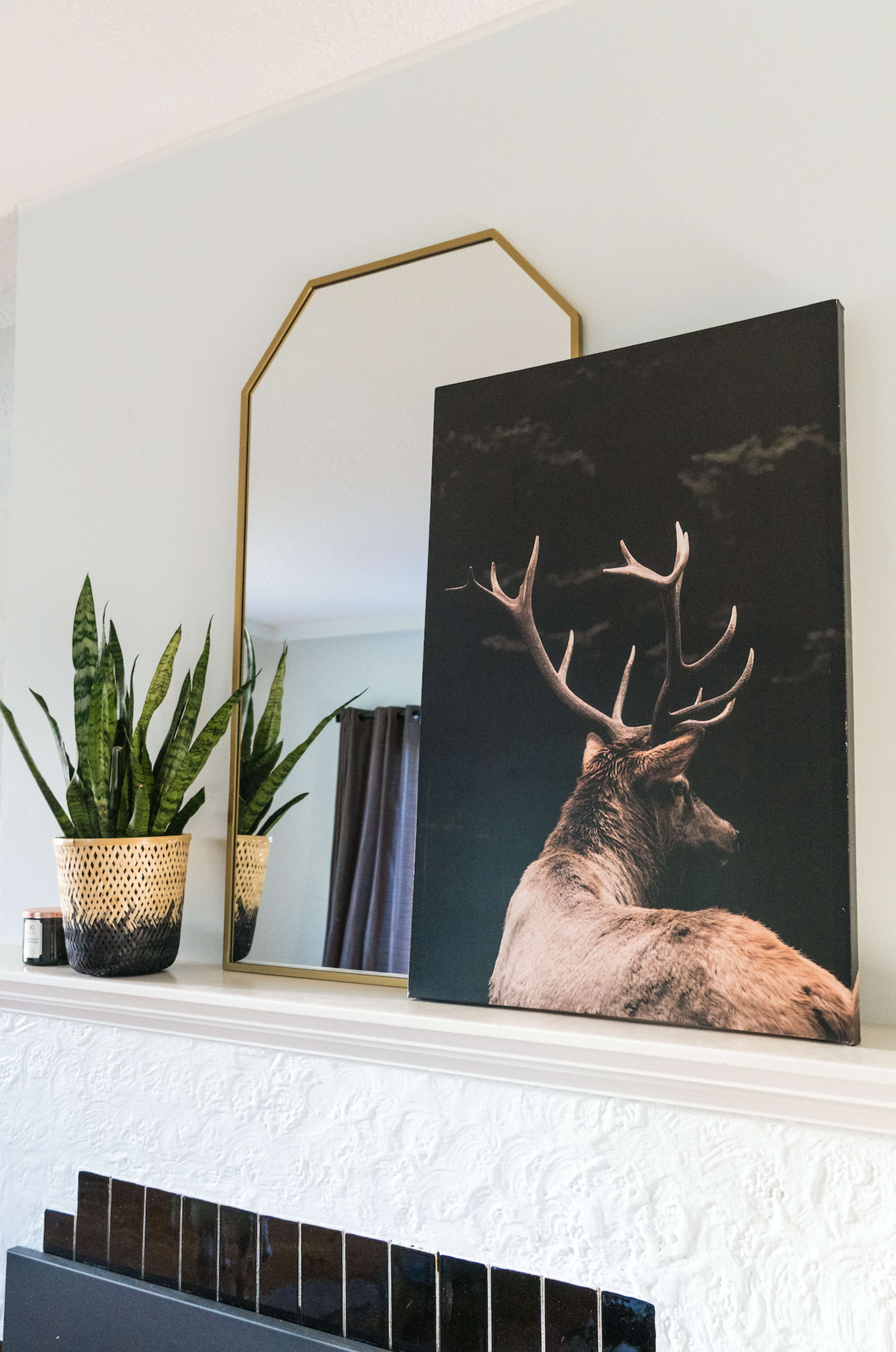 A mounted deer canvas is displayed above a fireplace, alongside a gold-framed mirror and potted plants.
