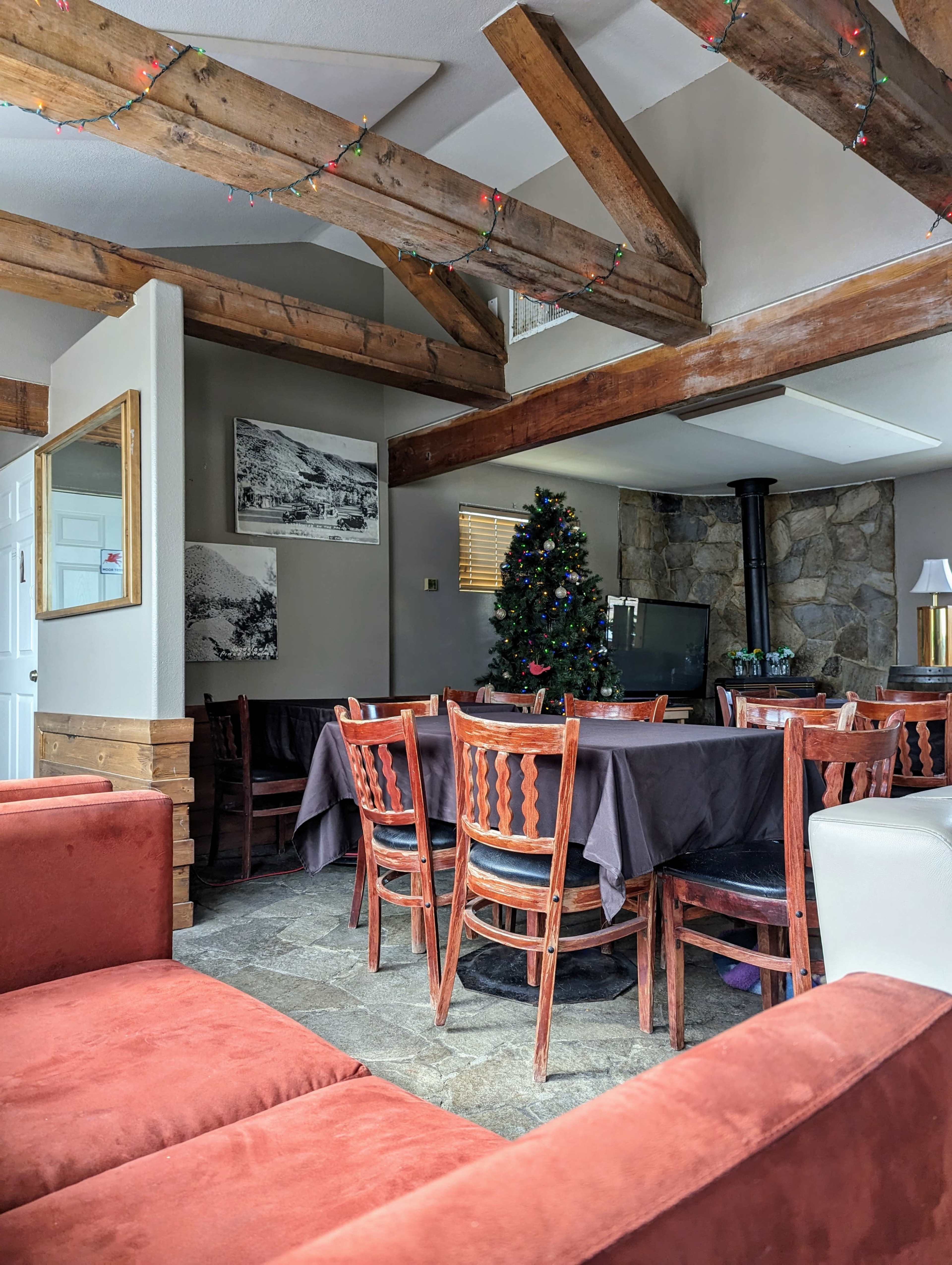 A dining area features a large table surrounded by wooden chairs, with a Christmas tree and a stone fireplace in the background.