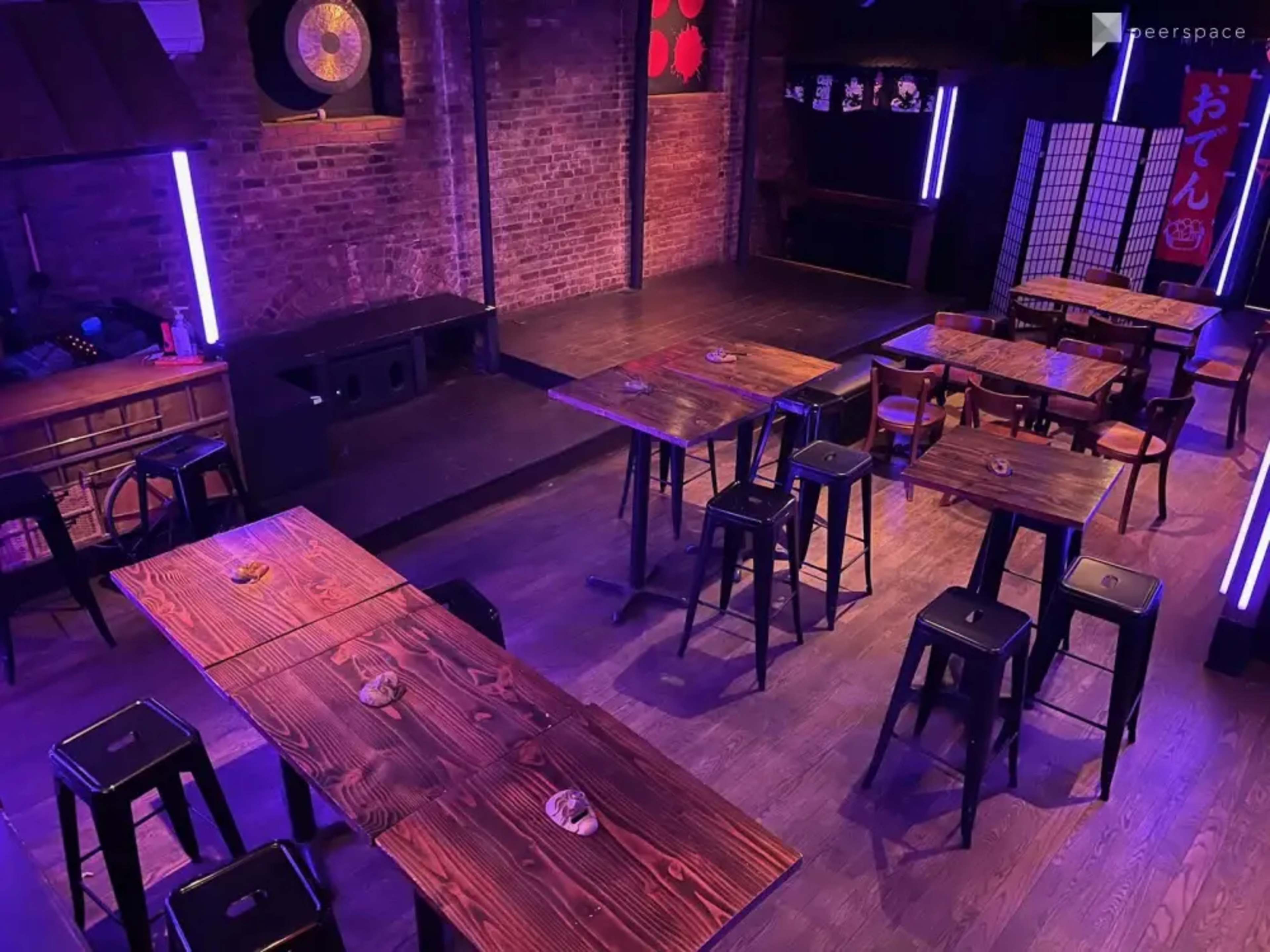 The image shows a dimly lit bar or restaurant interior with wooden tables and stools arranged in an open space, accented by purple LED lights and brick walls.