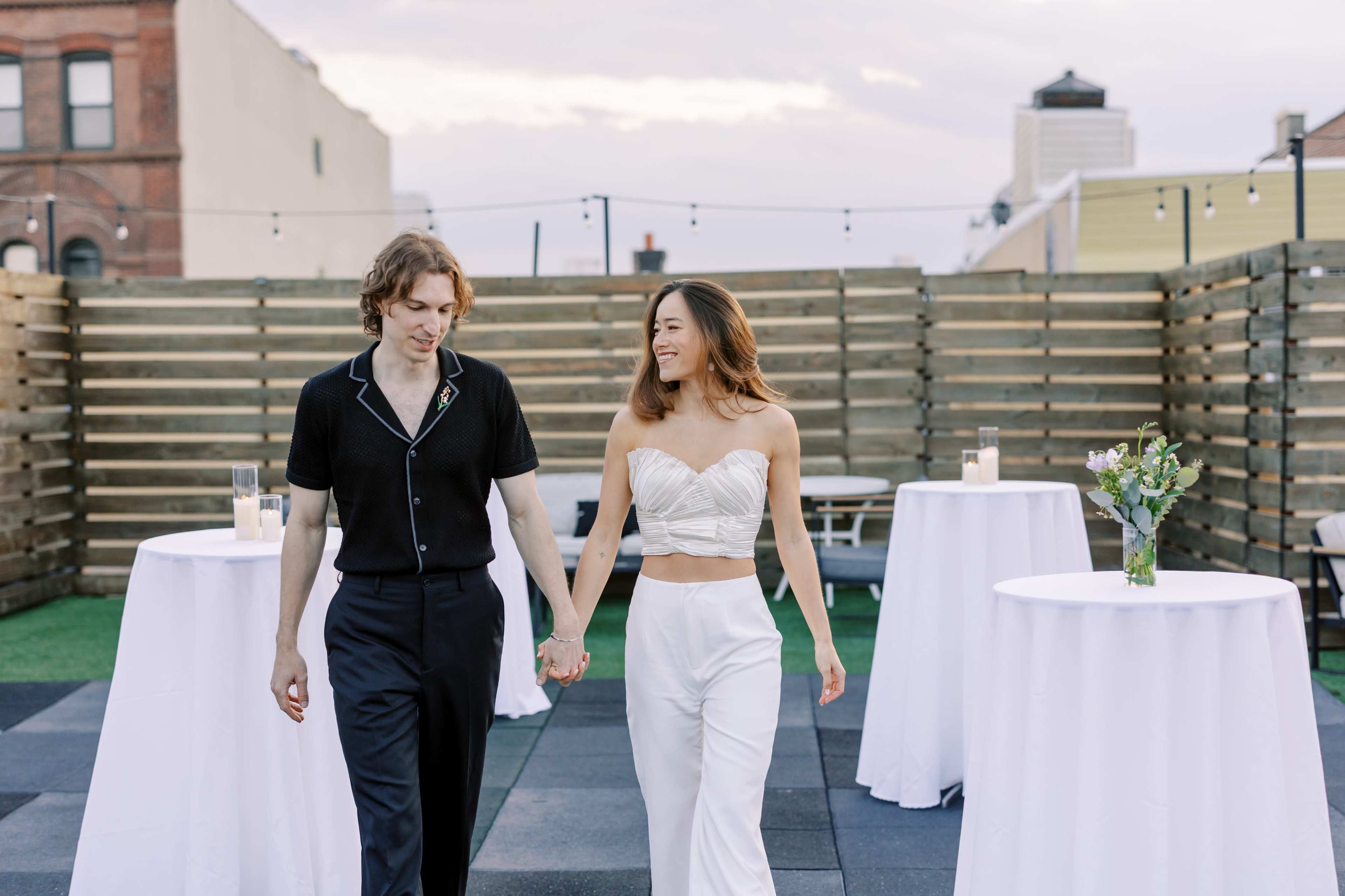 A couple walks hand in hand across a rooftop event space with white tables and a wooden fence in the background.